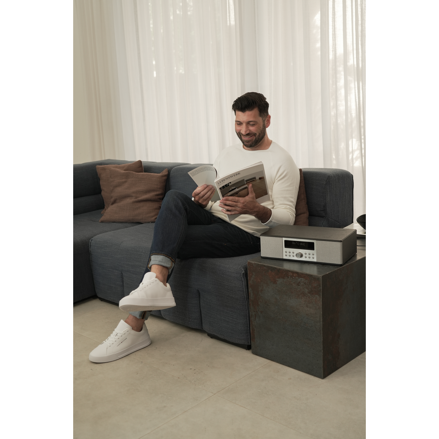 A man reading a magazine on a blue sofa next to a stereo.