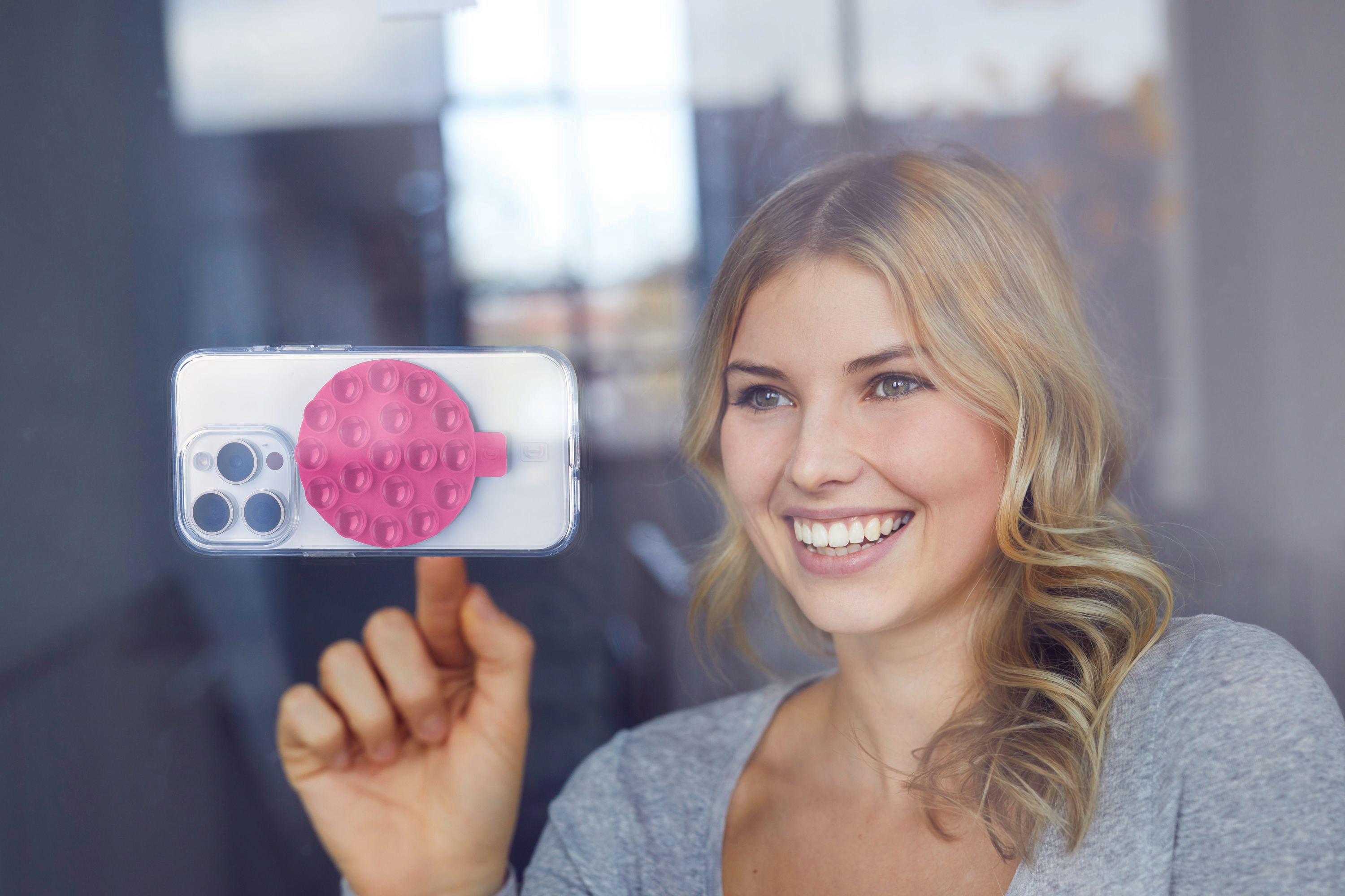 Eine Frau lächelt, während sie ein Telefon mit einem rosa Saugnapf an einem Fenster hält.