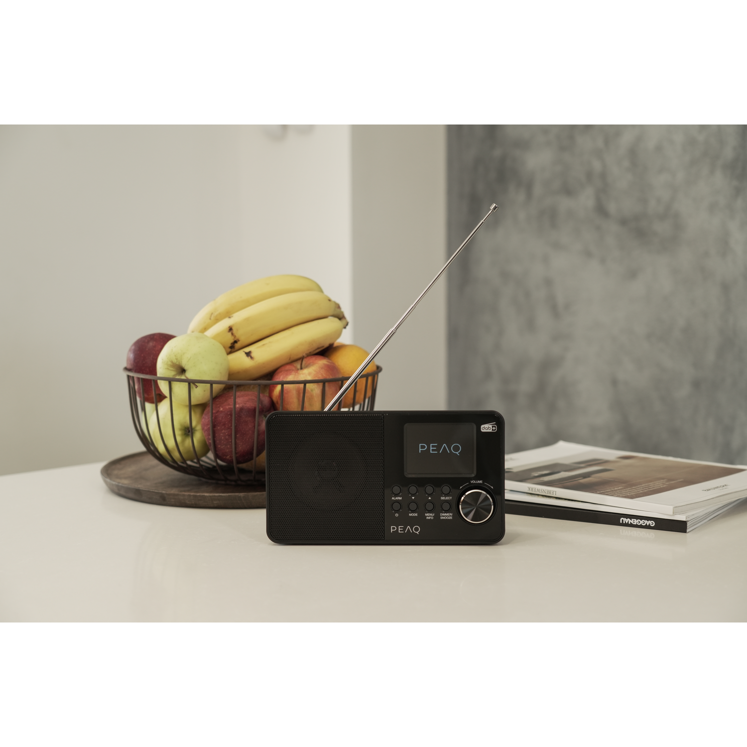 Black radio with antenna on white table, fruit basket, and books. Background includes a wall.