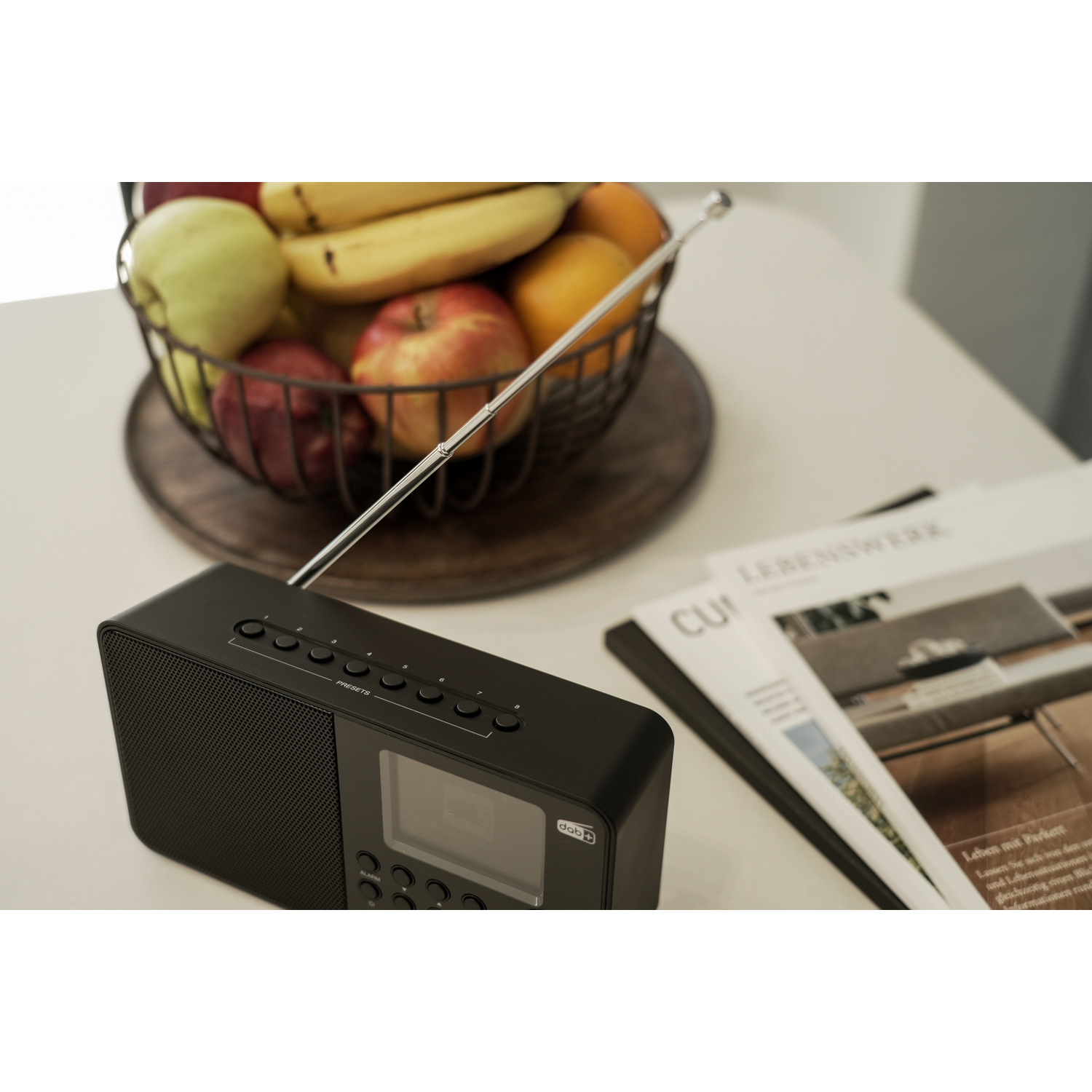 Black radio with a display and fruits in a basket on a white table. Magazine in the background.