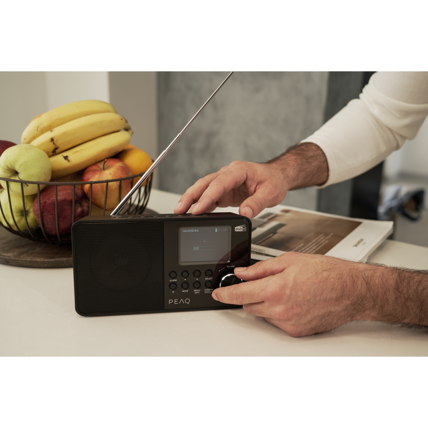 A person adjusts the dial on a black radio, beside a fruit basket.