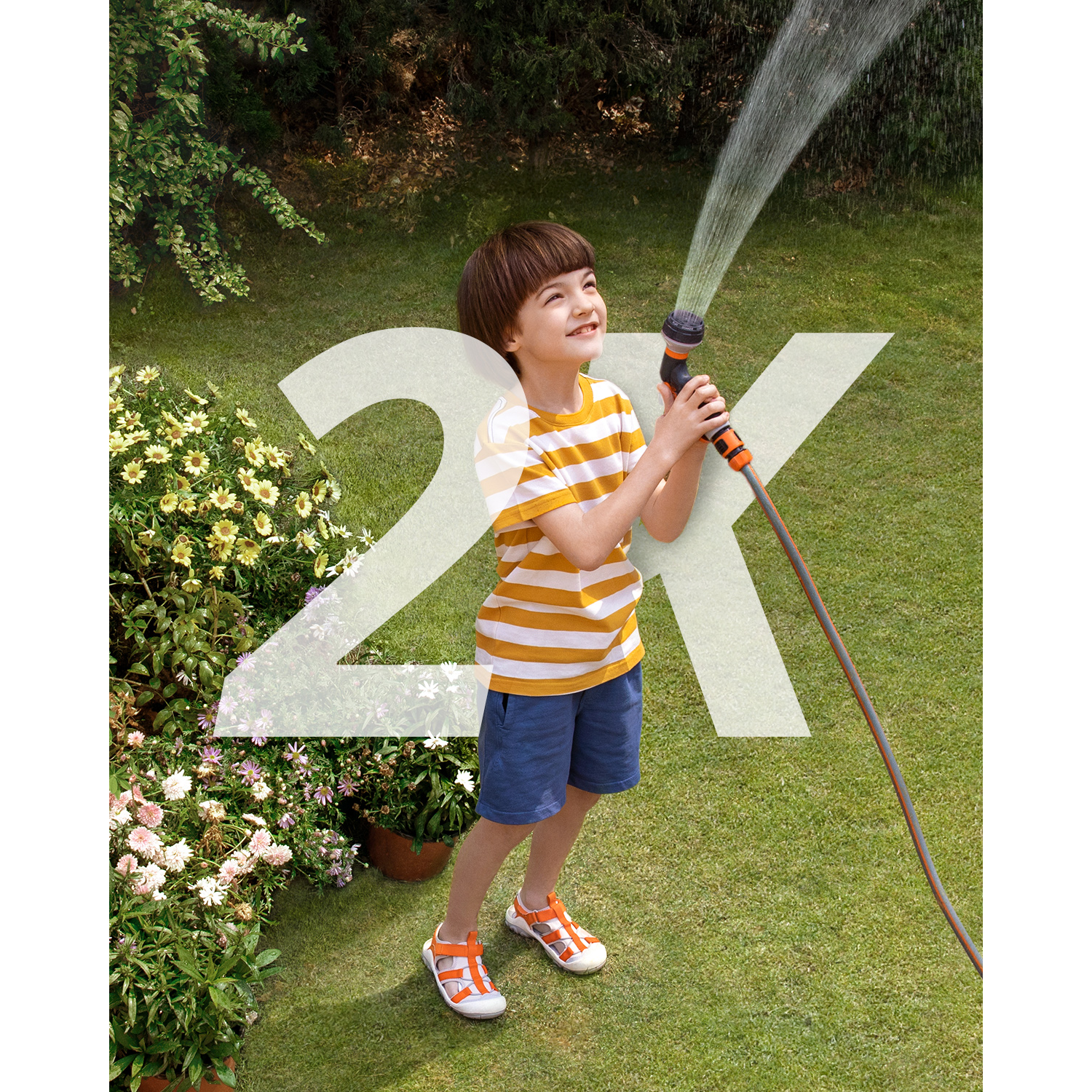 A young boy is holding a hose and spraying water in a garden, with a large 2X overlay.