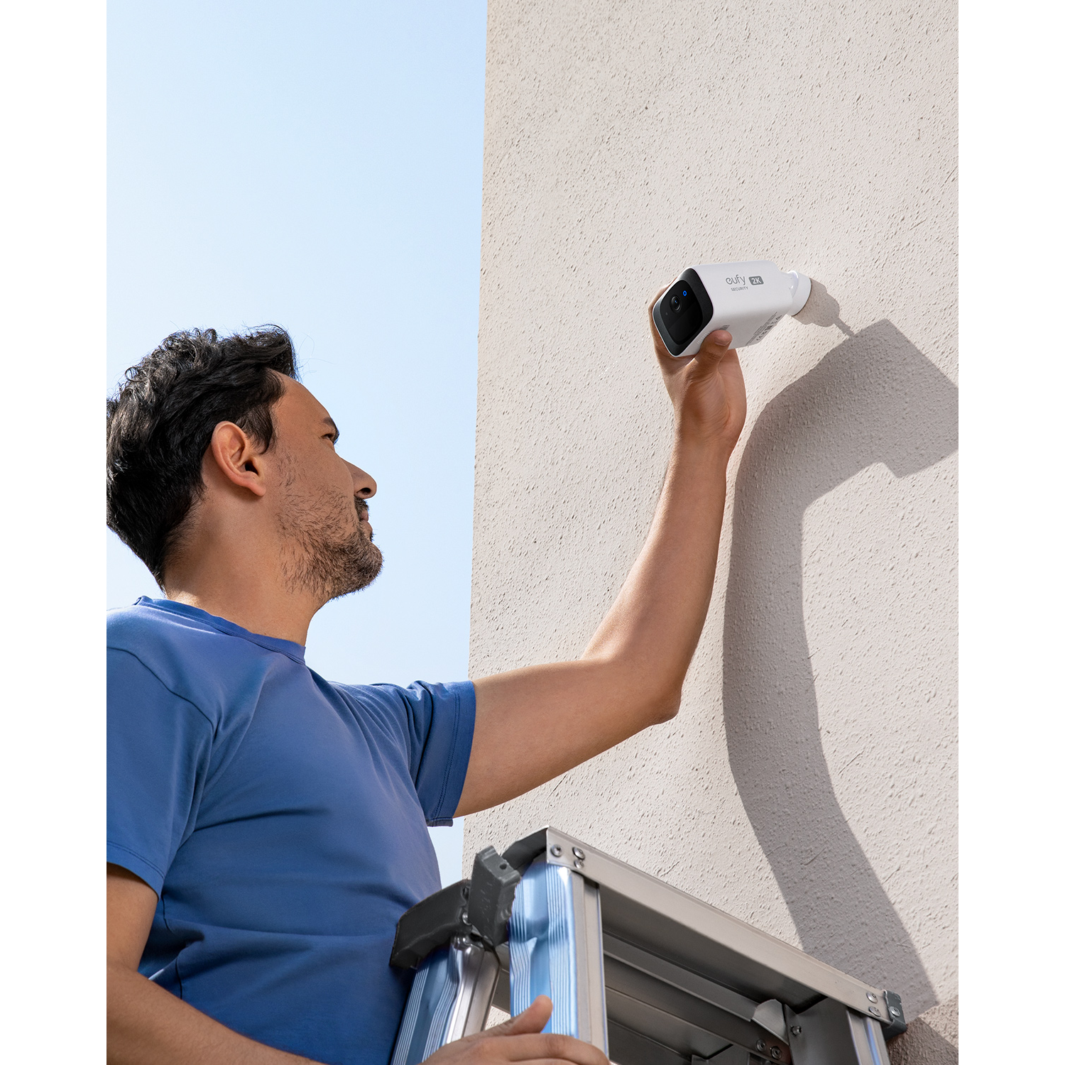 A man on a ladder installing a white outdoor security camera on a wall.