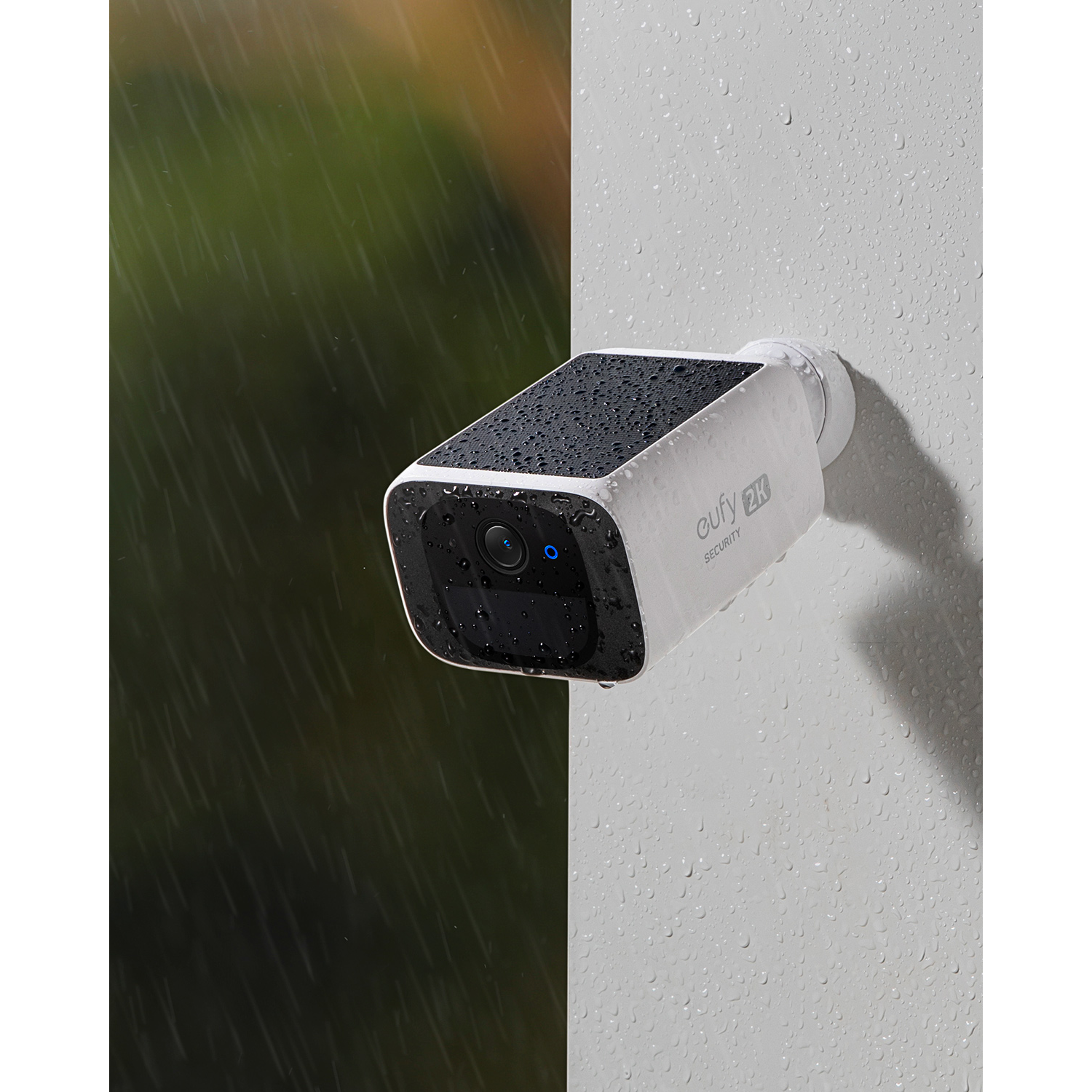 A white security camera on a wall with water droplets. It is raining.