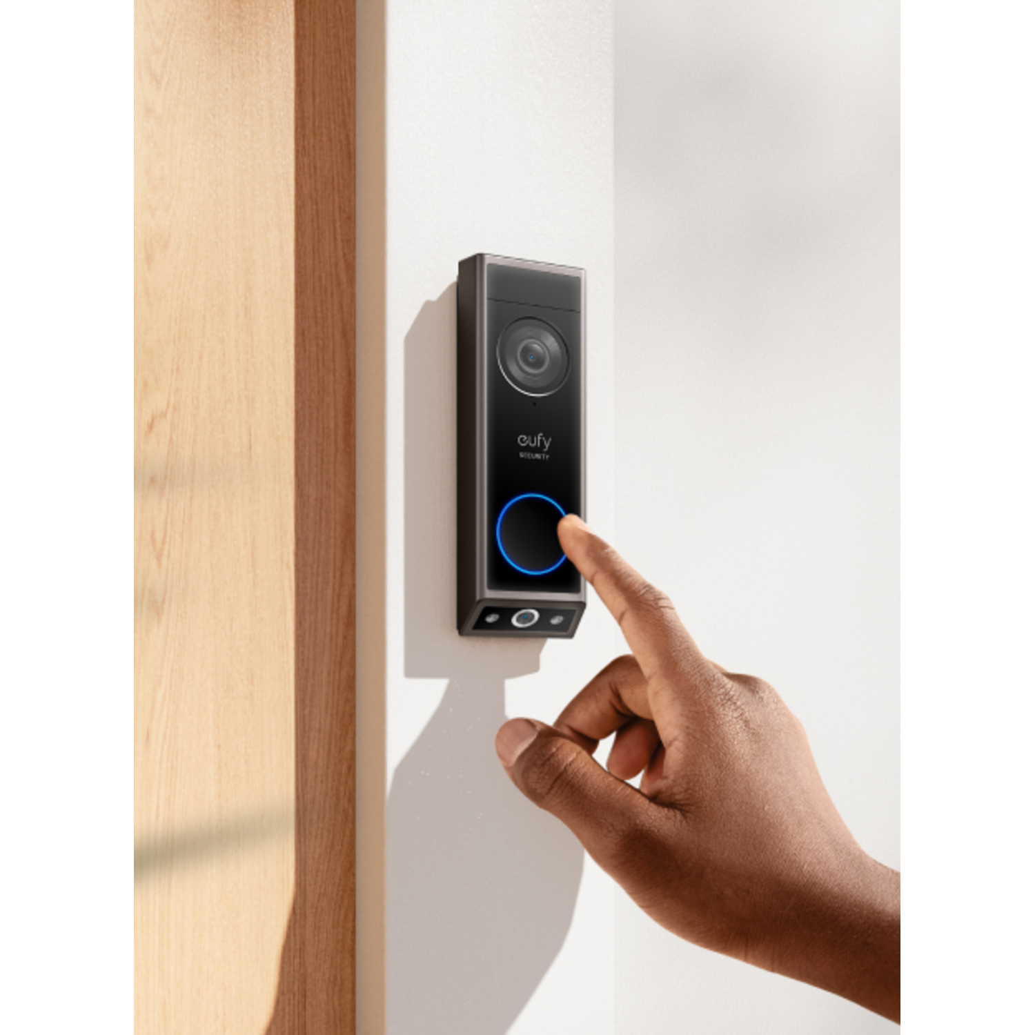 A hand presses a doorbell button on a black device mounted on a white wall.