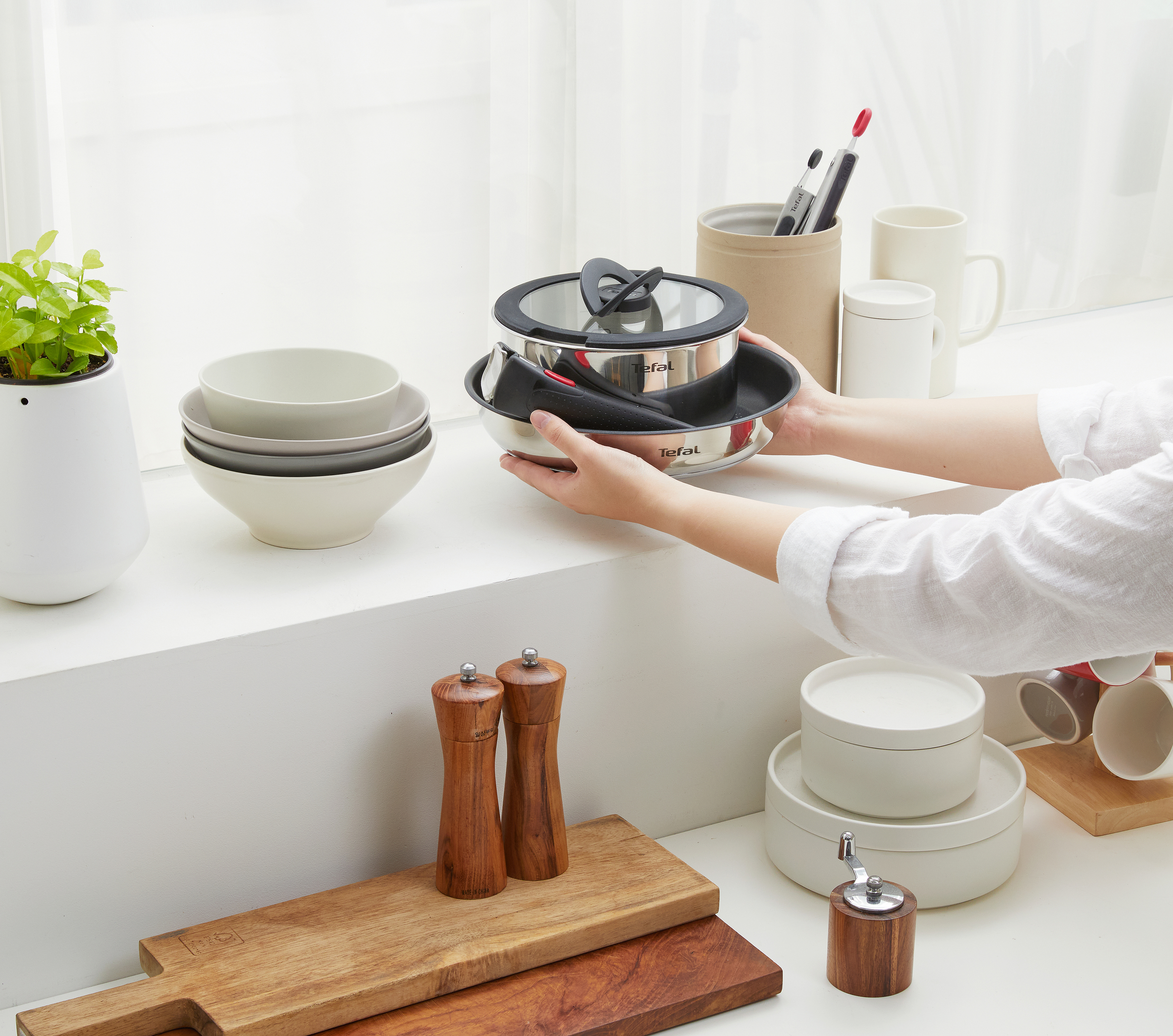Osoba trzymająca garnek Tefal z innymi przedmiotami kuchennymi na białej powierzchni.