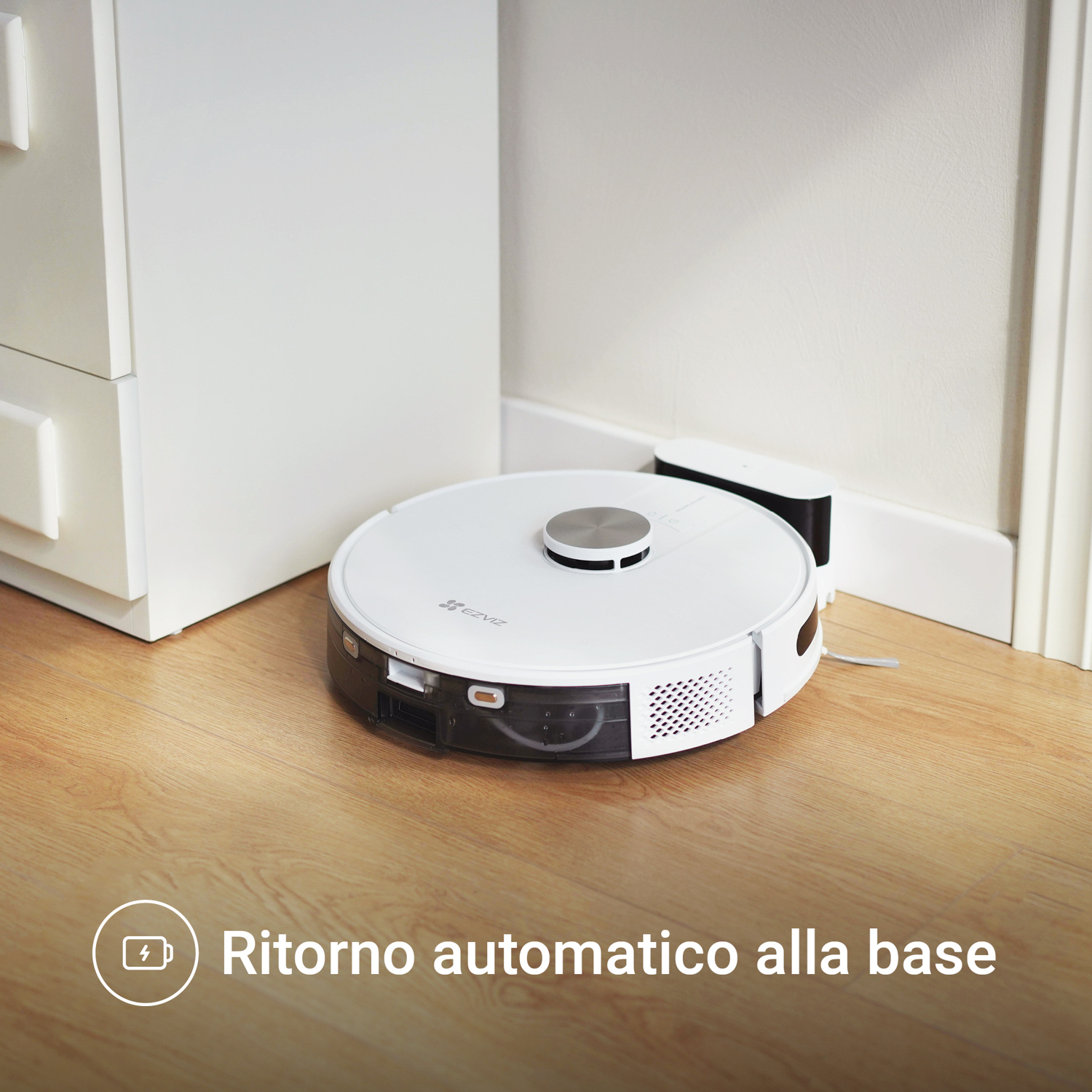 Un aspirapolvere robotico bianco su un pavimento in legno. Ha una stazione di ricarica. Il testo dice Ritorno automatico alla base.