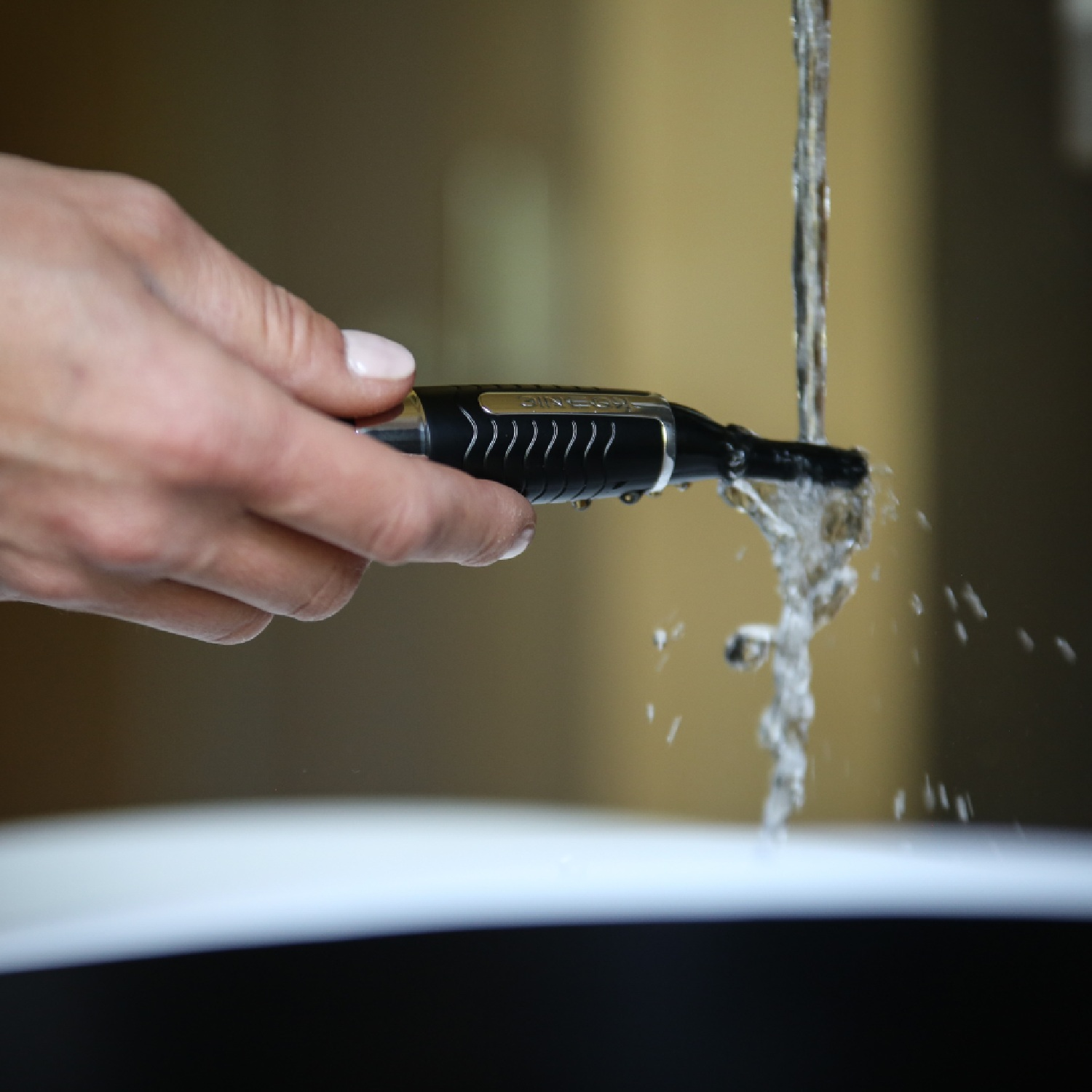 Een hand die een zwarte trimmer onder stromend water houdt. Water stroomt van bovenaf, wazig.