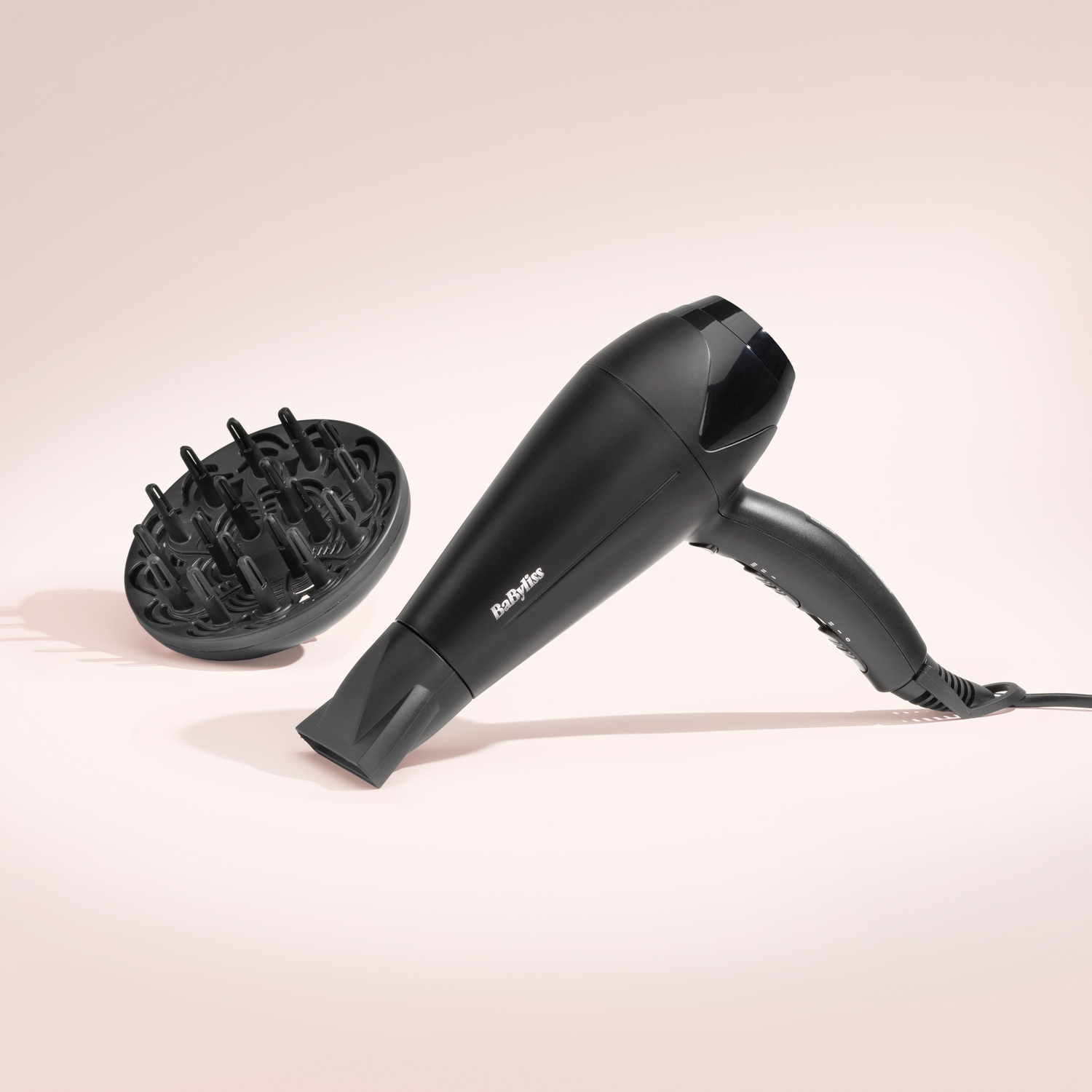 Secador de pelo negro y difusor sobre fondo rosa. La marca 'Babyliss' es visible.