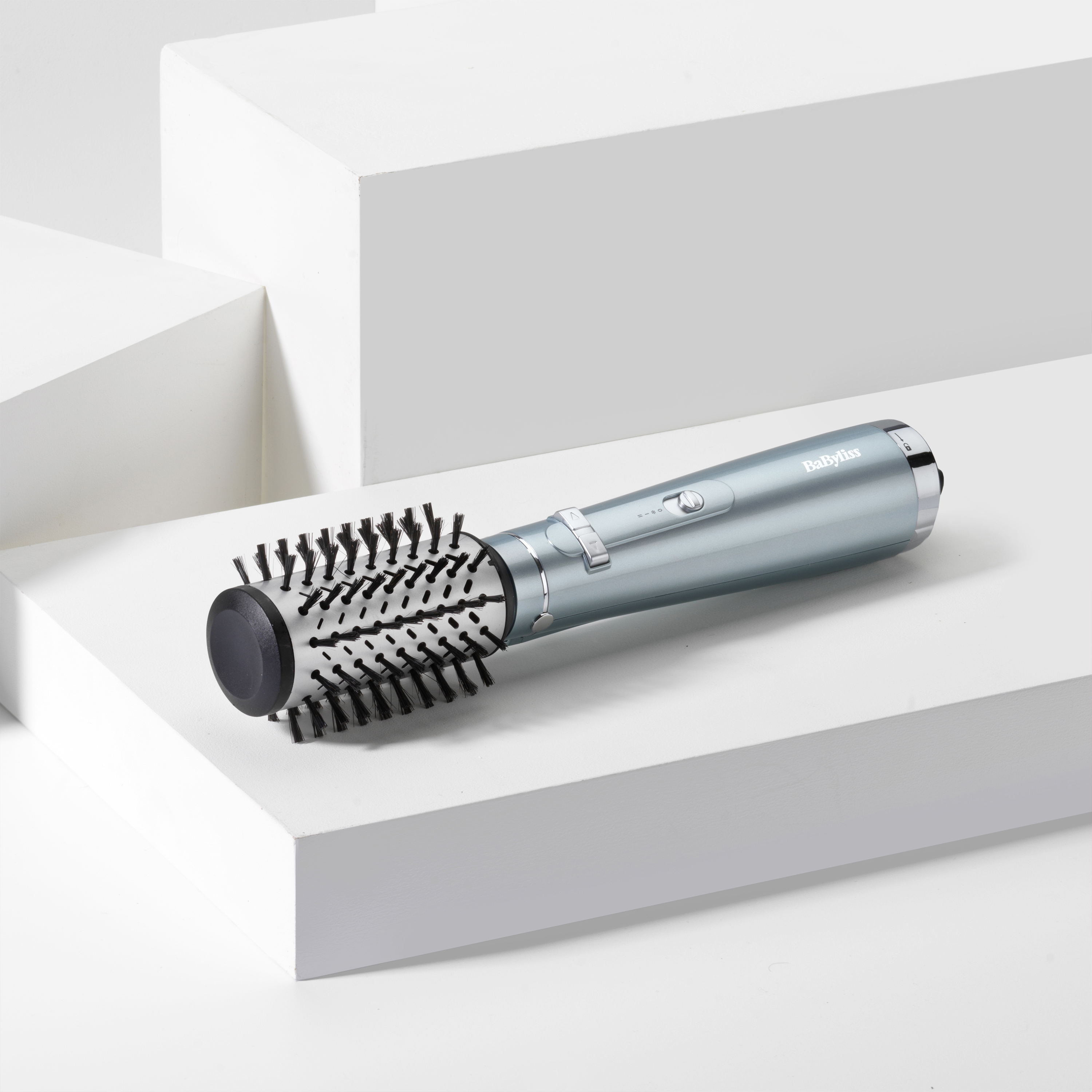 Un moldeador de pelo color turquesa sobre bloques blancos. Tiene un cabezal de cepillo y el logo de Babyliss.