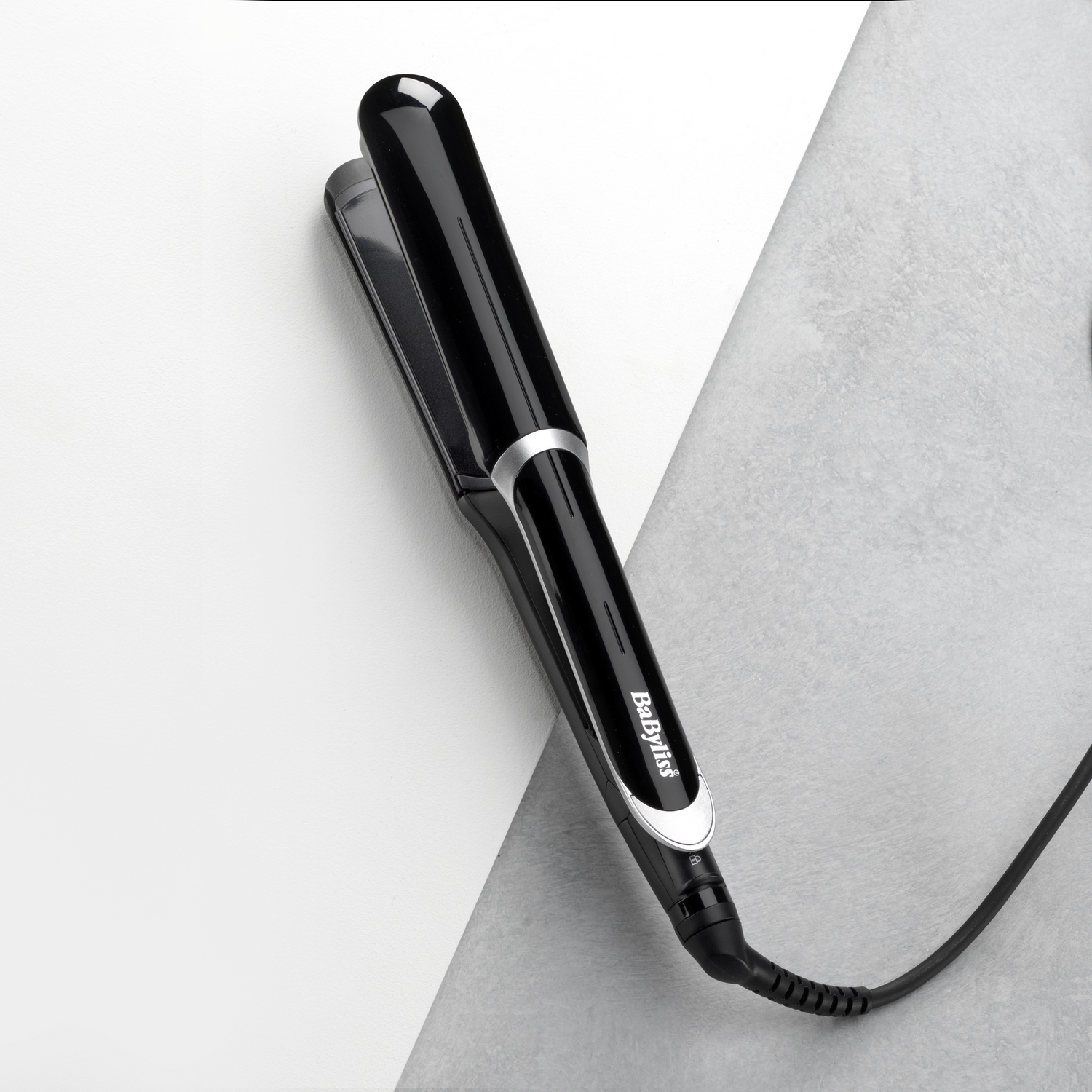 Alisador de pelo negro sobre una superficie blanca y gris. Texto: BaByliss.