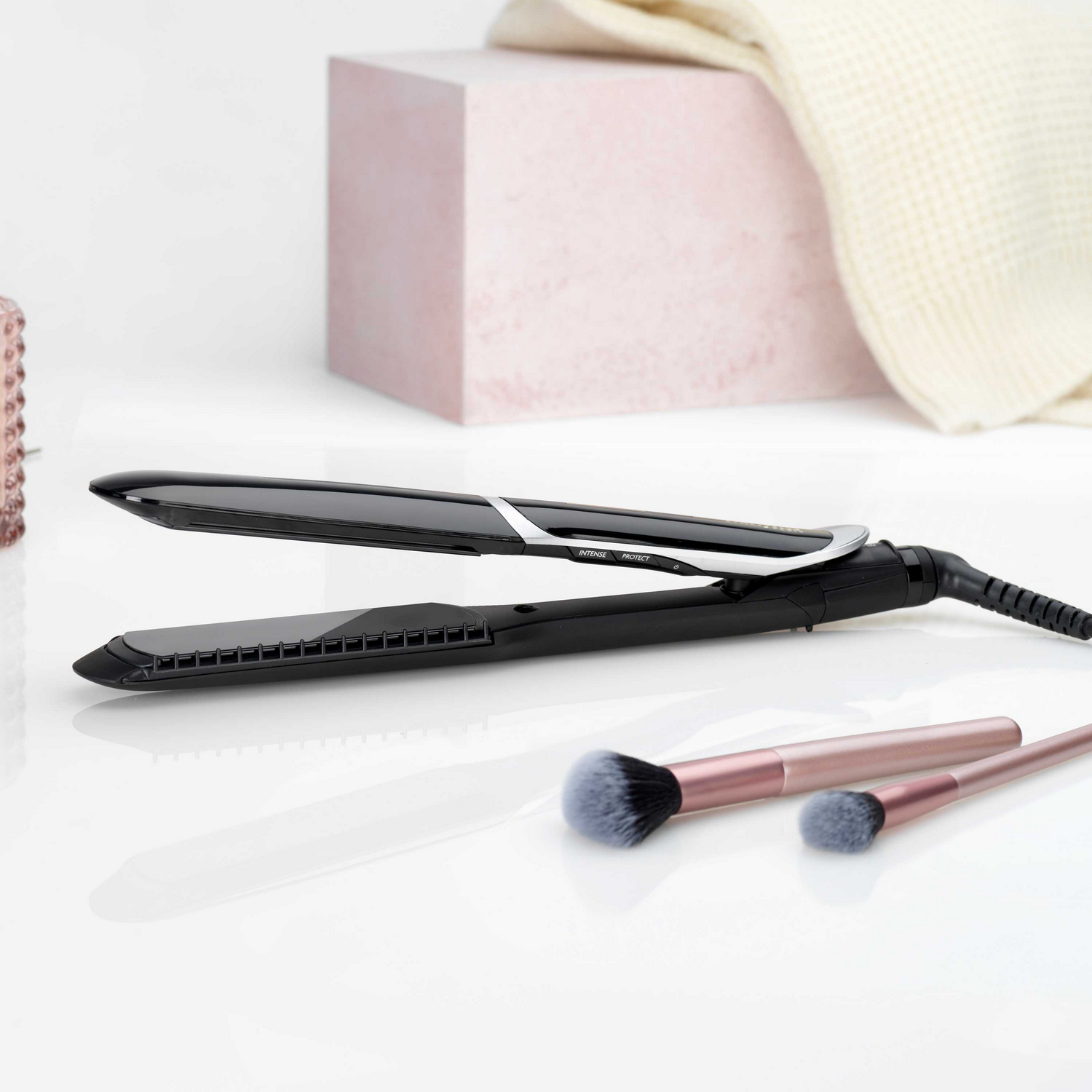 Plancha de pelo negra y dos brochas de maquillaje sobre una superficie blanca, caja rosa detrás.