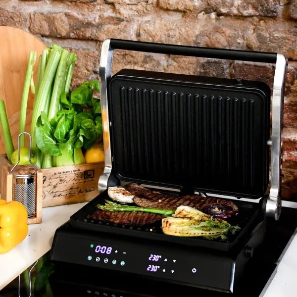 Otwarty grill z ugotowanym jedzeniem, obok warzyw na blacie z tłem z ceglanej ściany.