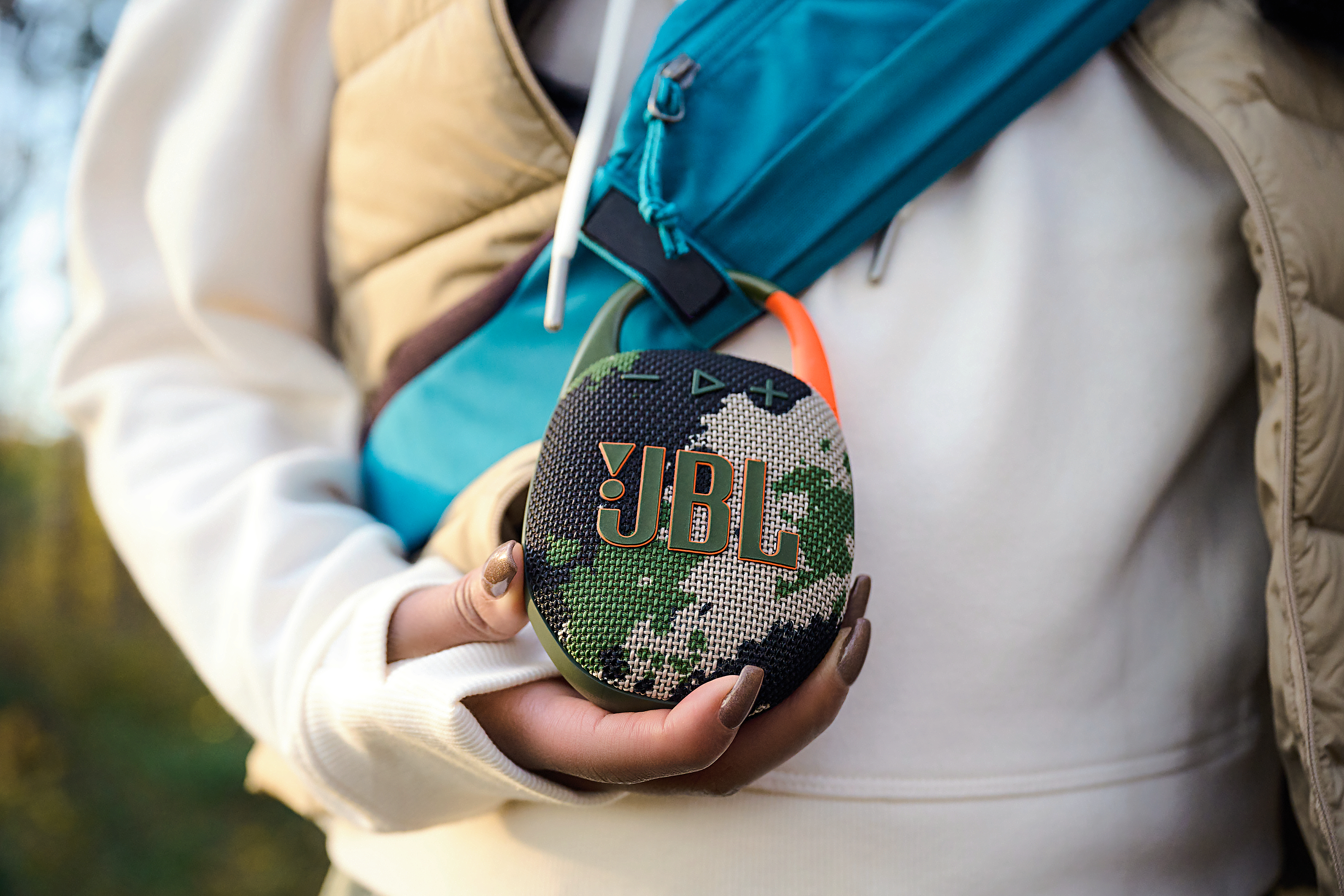 Eine Hand, die einen Camo-JBL-Lautsprecher mit einer blauen Bauchtasche und Weste hält.