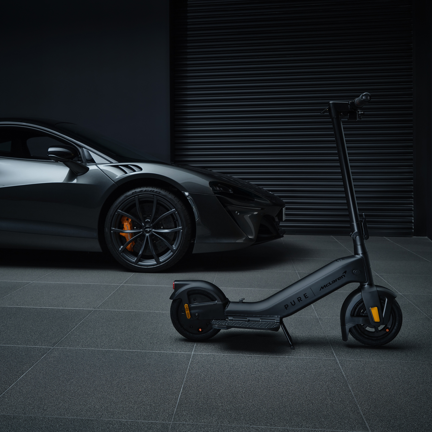 Black McLaren scooter and a gray McLaren car, in a dark setting with a textured wall.