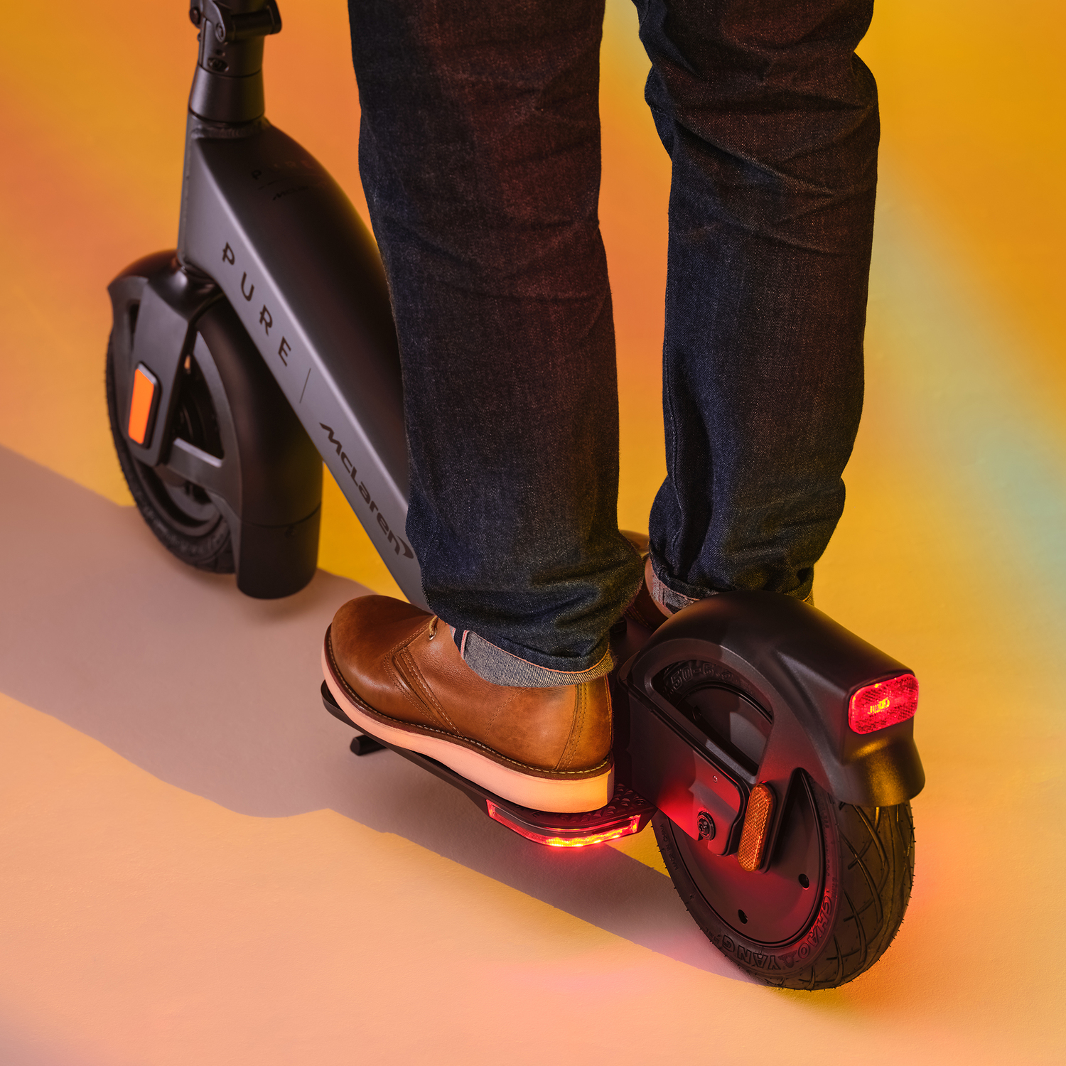 Person riding a black and gray Pure McLaren electric scooter, with jeans and brown shoes.