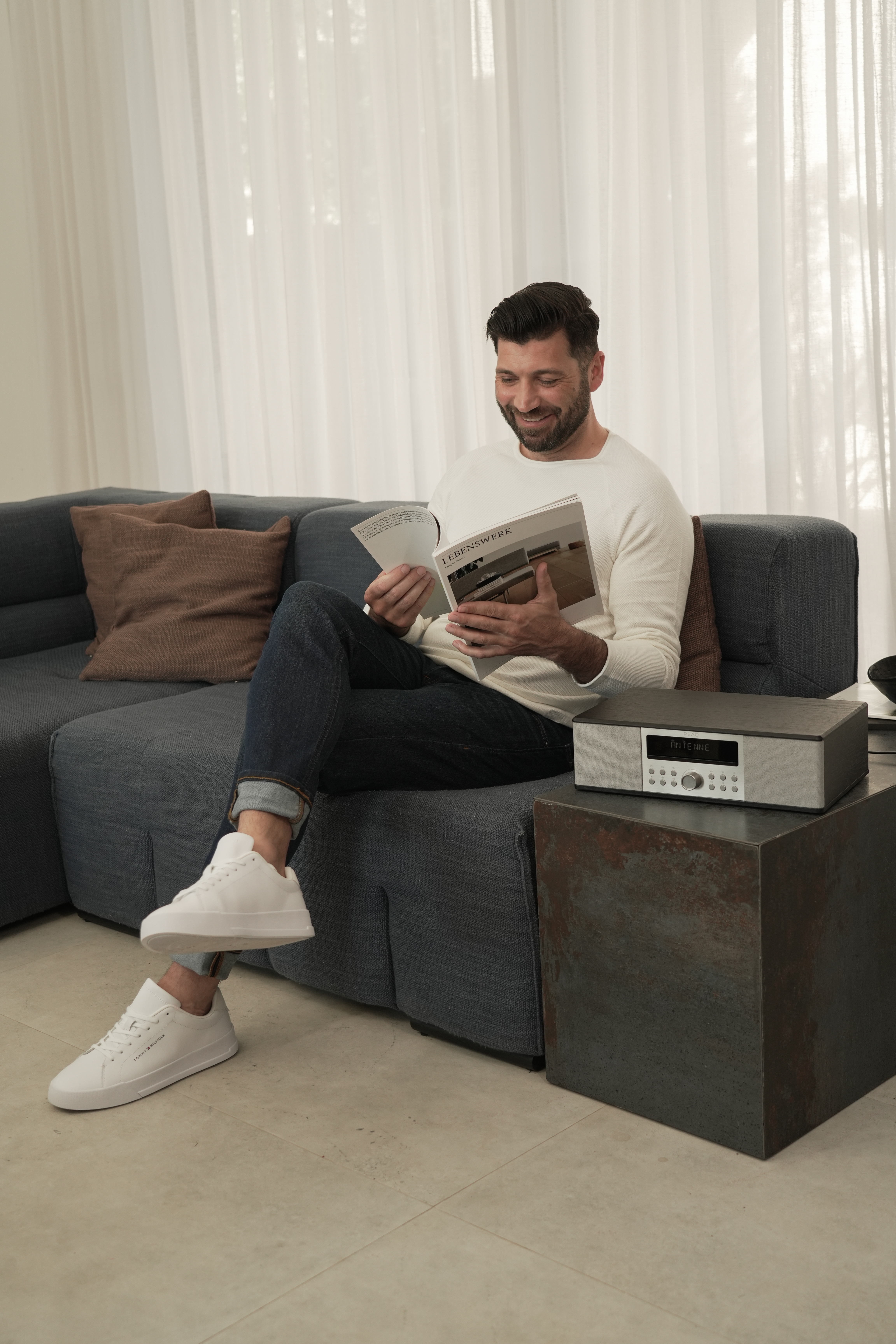 Un homme est assis sur un canapé, lisant un magazine. Une radio et une table d'appoint sont à côté de lui. La pièce a un design moderne.