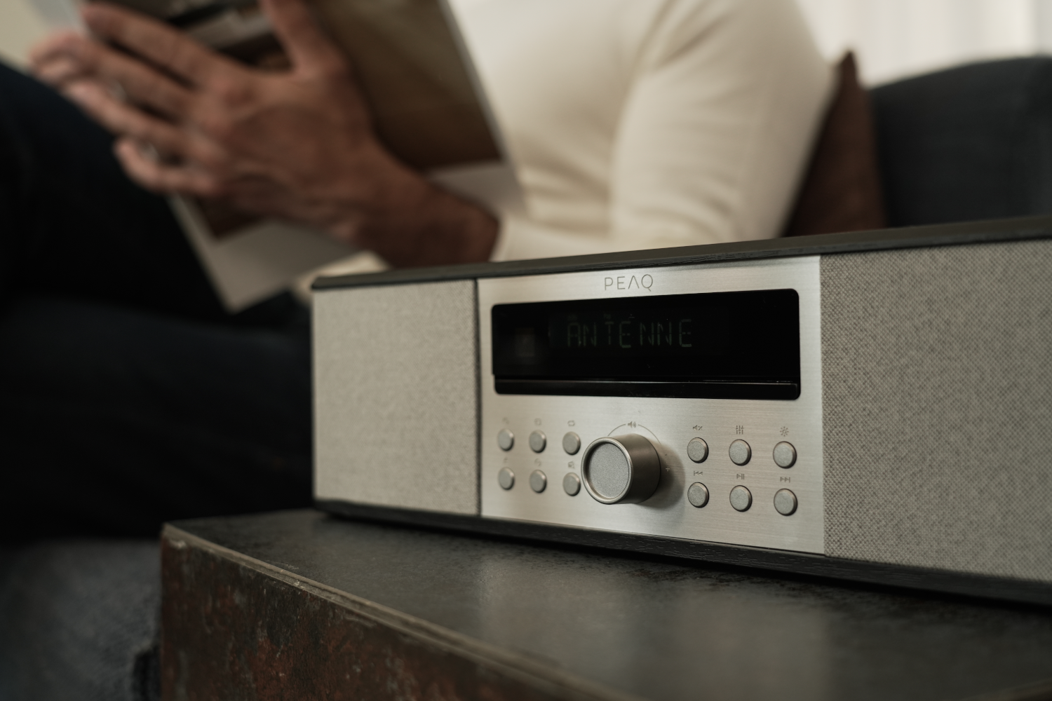 Une radio avec un écran indiquant 'Antenne'. Une personne lit un livre en arrière-plan.