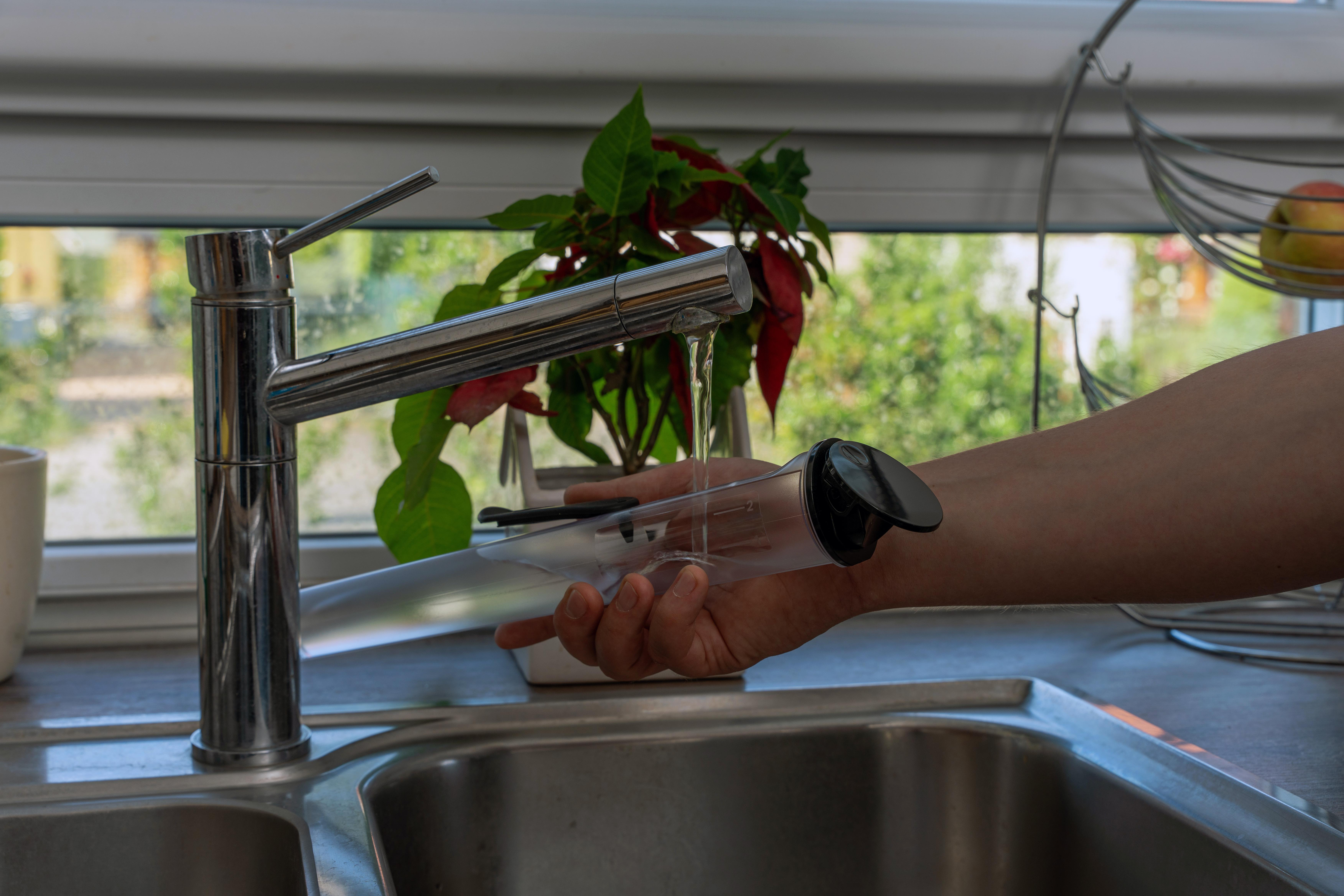 Eine Hand hält einen durchsichtigen Behälter unter einen Wasserhahn. Wasser fließt in den Behälter.