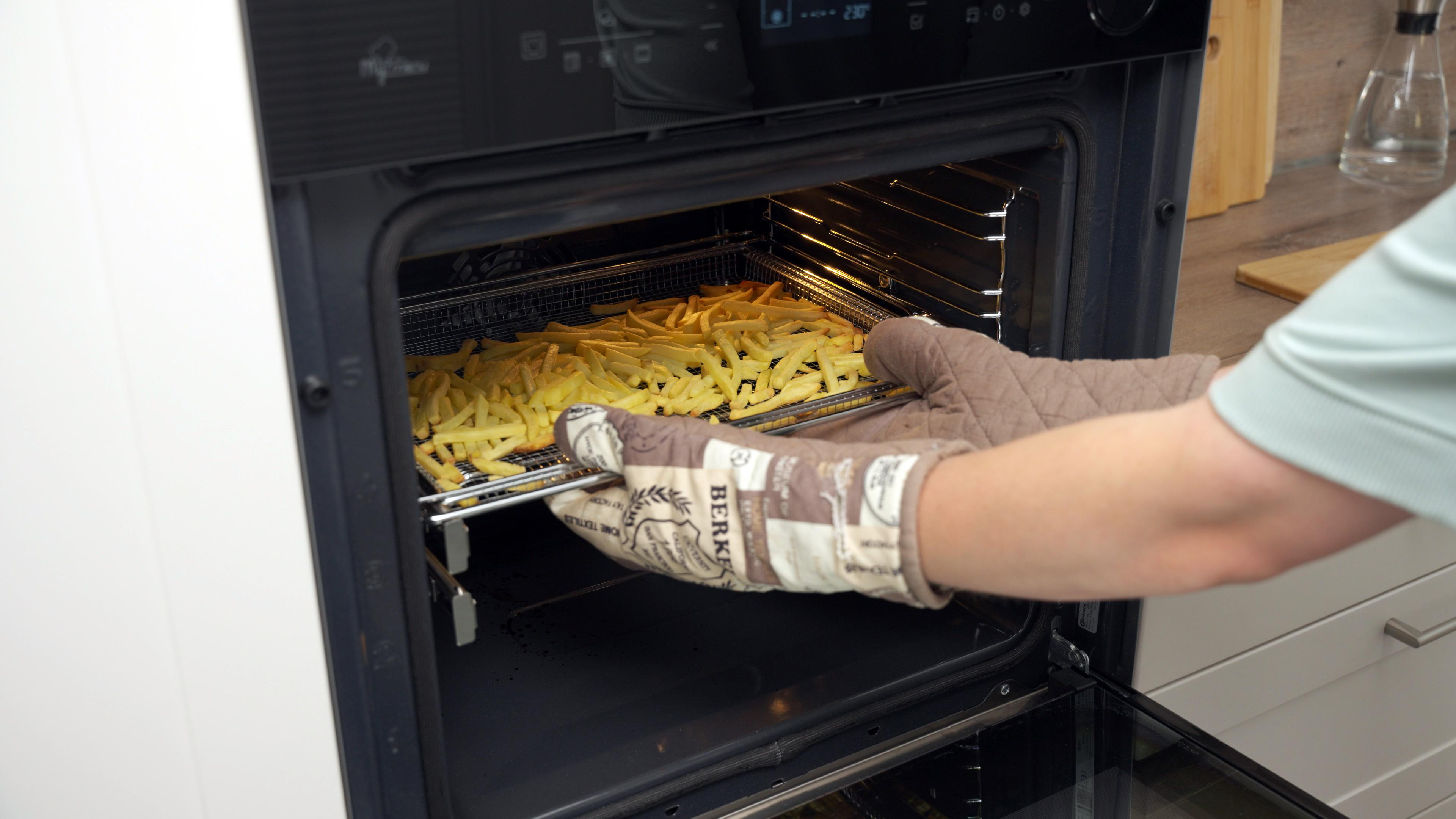 Eine Person entnimmt ein Blech mit Pommes Frites aus dem Ofen, mit einem Ofenhandschuh.