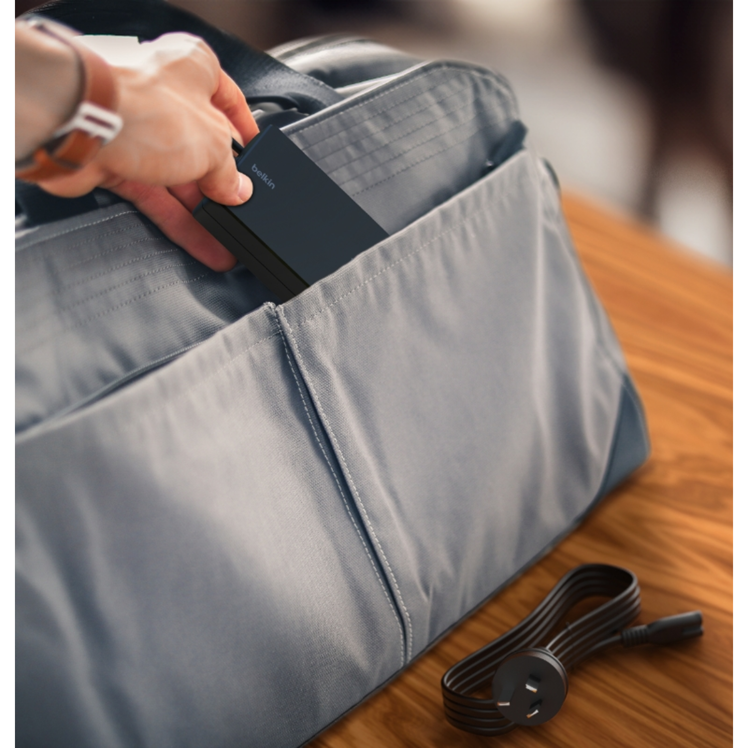 A hand putting a black power bank into a gray bag. A cable is also visible.