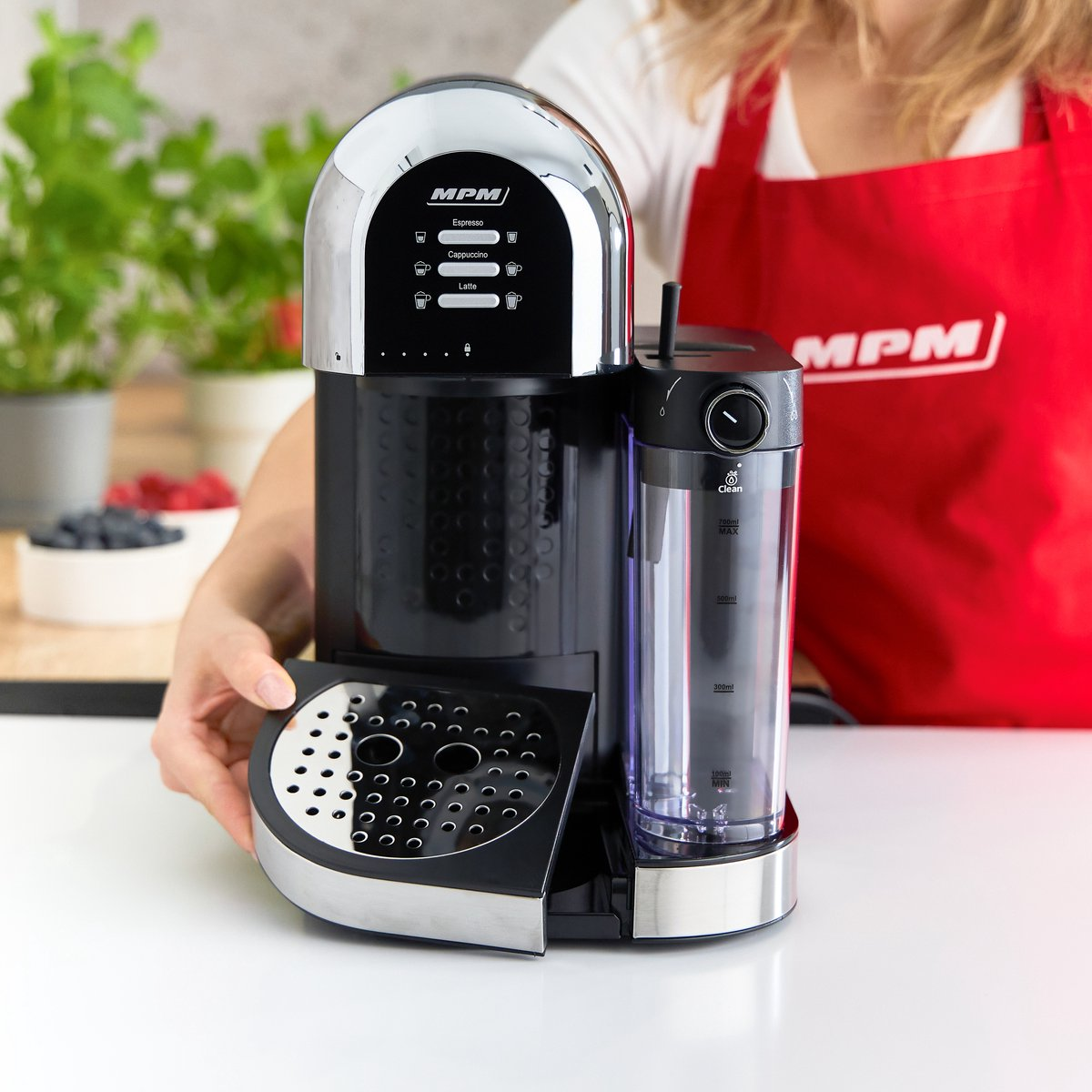 Osoba trzyma czarno-srebrny ekspres do kawy, czerwony fartuch w tle, ustawienie kuchenne.