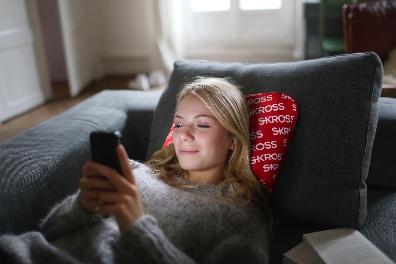 Blonde Frau liegt auf einem grauen Sofa und benutzt ein Telefon. Rotes Kissen hinter ihr mit SKROSS darauf.