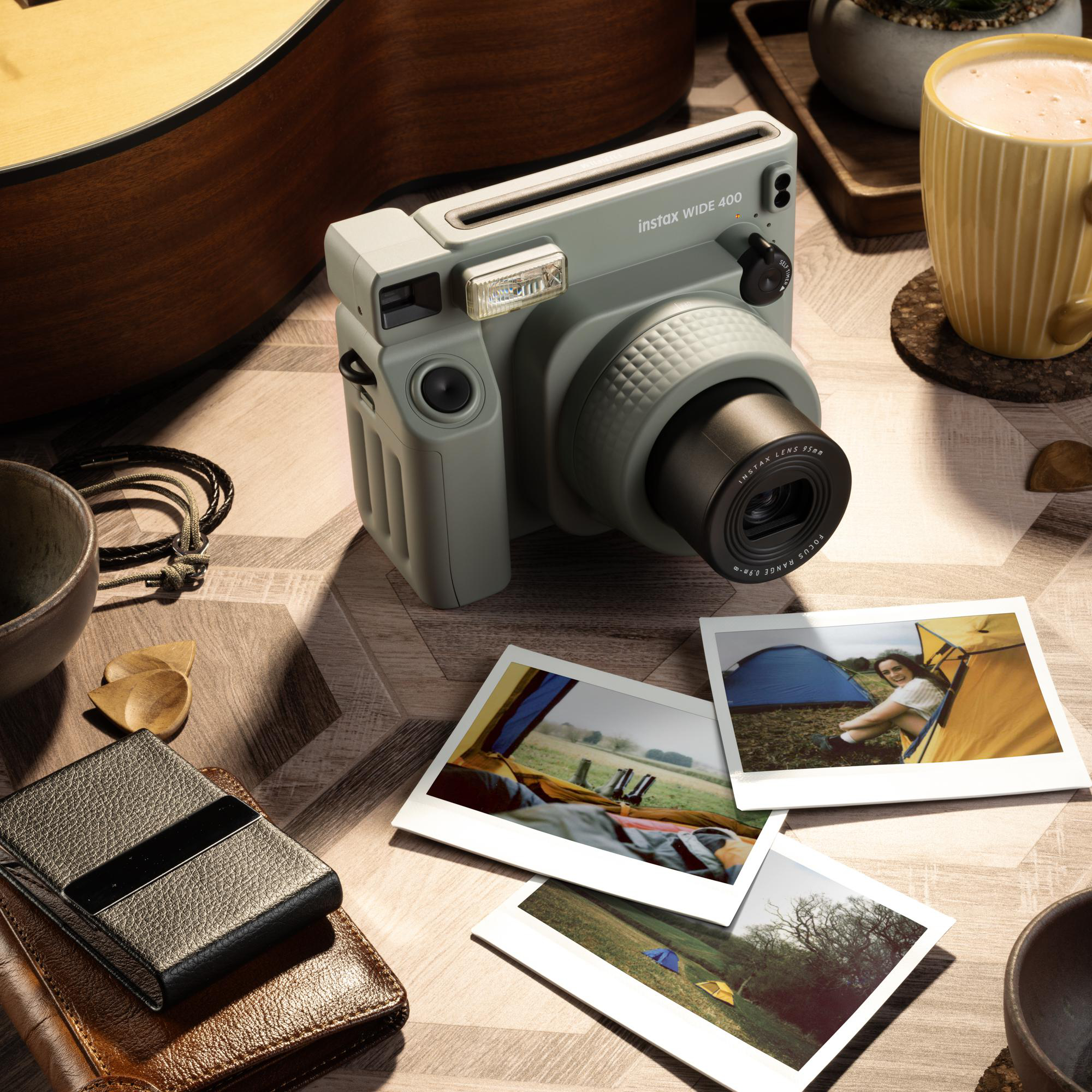 Graue Kamera, Fotos und eine Tasse auf einem Holztisch. Im Hintergrund eine Gitarre.