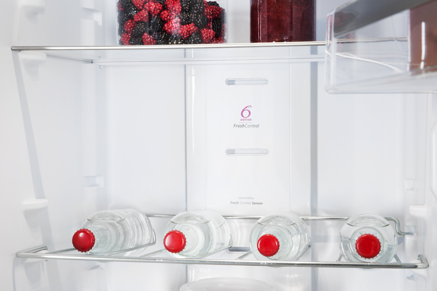 Dentro de un refrigerador. Botellas de vidrio con tapas rojas están en un estante. Bayas arriba.