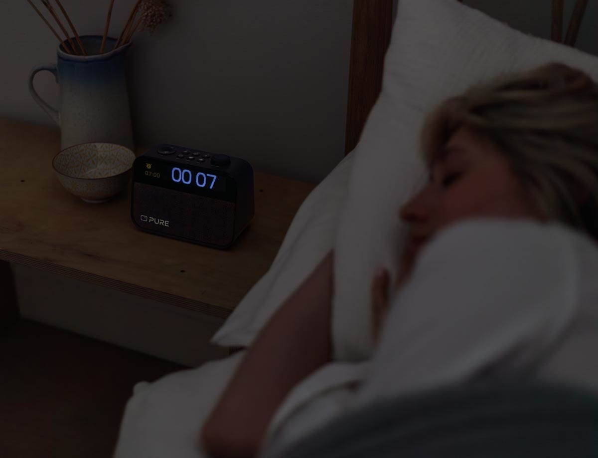 Une femme endormie dans son lit. Une horloge affichant l'heure '00:07' est sur la table de chevet à côté d'elle.