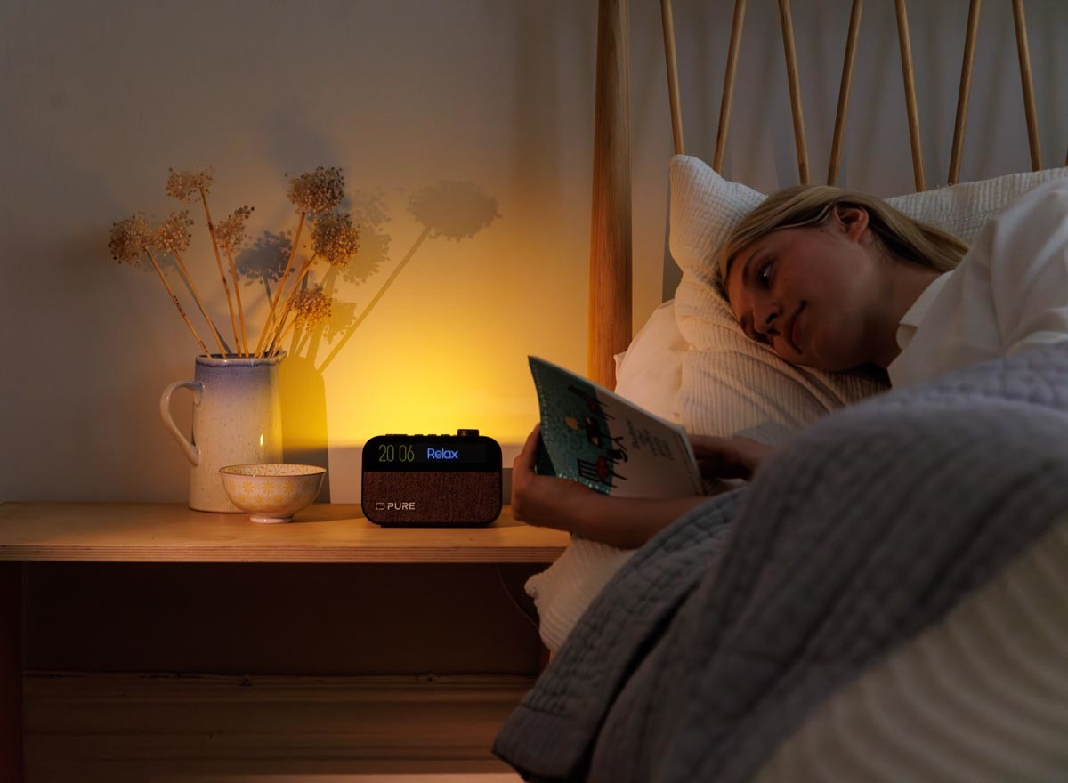 Scène de chambre à coucher la nuit. Femme au lit lisant avec une radio Pure sur une table.