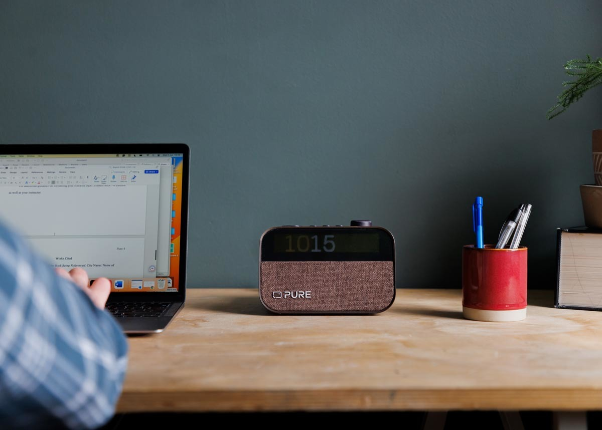 Bureau avec ordinateur portable, radio, porte-stylos et bras d'une personne en arrière-plan.