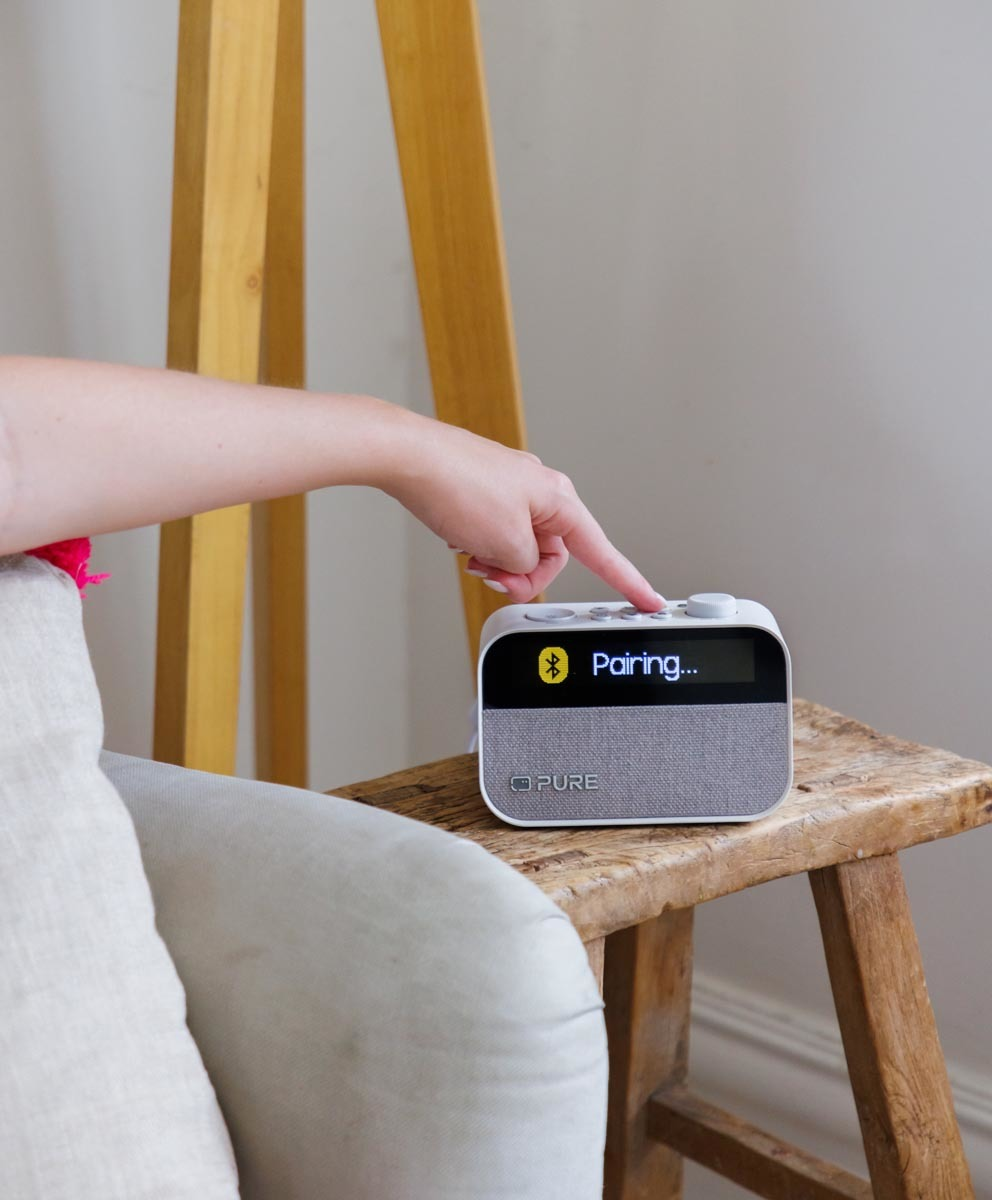 La main d'une personne appuie sur le bouton d'appairage d'une enceinte blanche et grise, sur un tabouret en bois.
