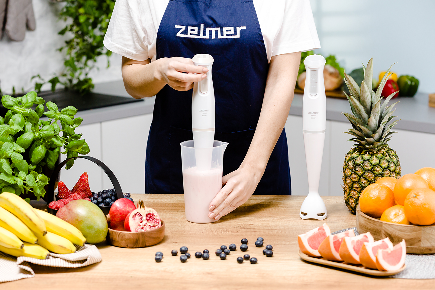 Nő rózsaszín turmixot kever egy Zelmer botmixerrel; gyümölcsök az asztalon.