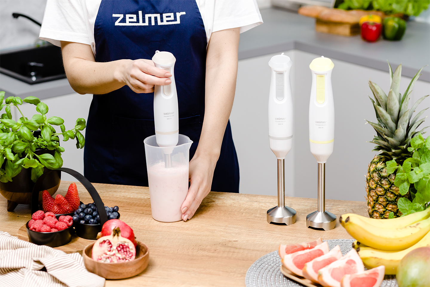 Személy turmixgépet használ, két másik turmixgéppel és gyümölcsökkel, faasztalon.