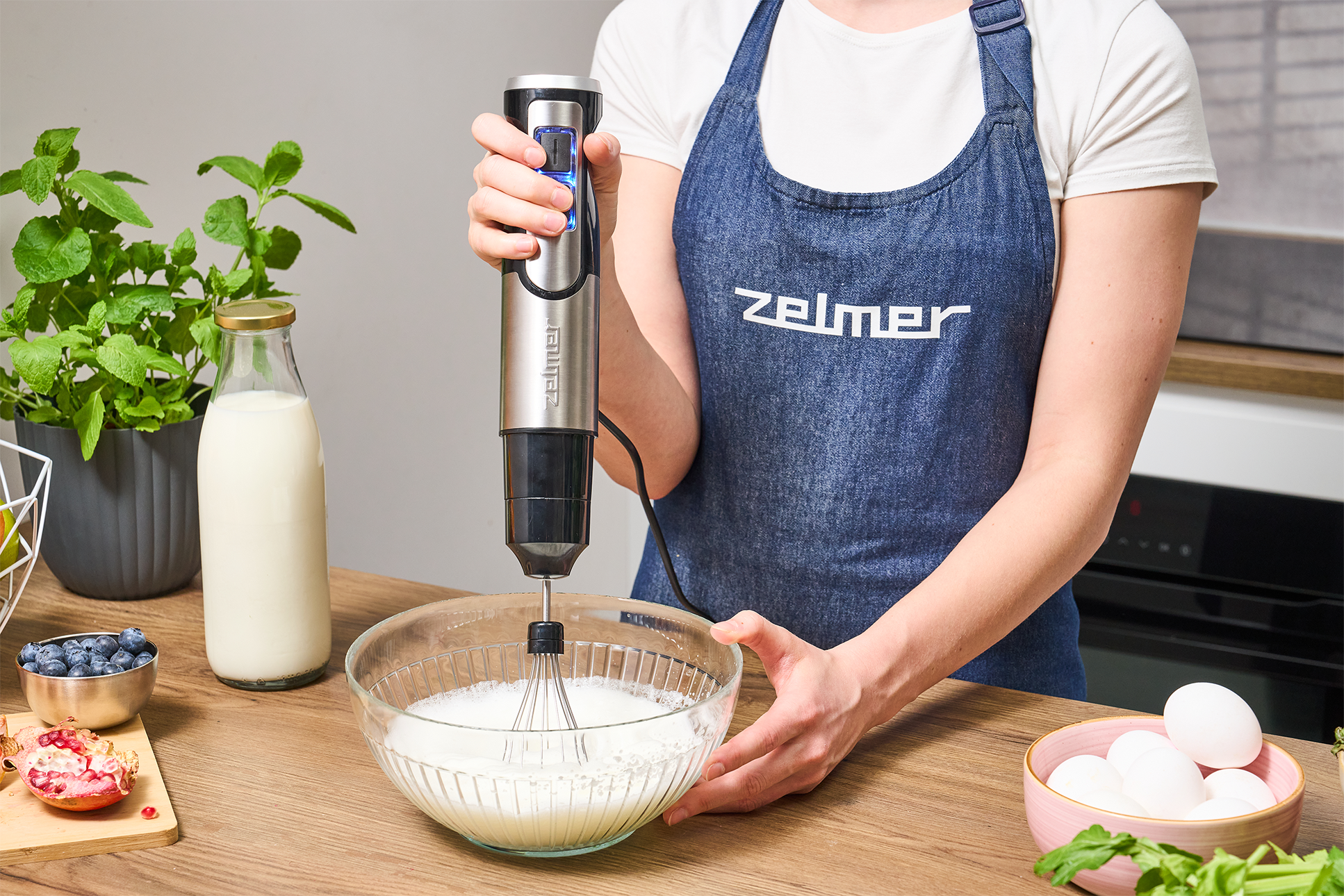 Egy kék kötényes nő kézi turmixgéppel keveri az összetevőket egy üvegtálban. Konyhai környezet.