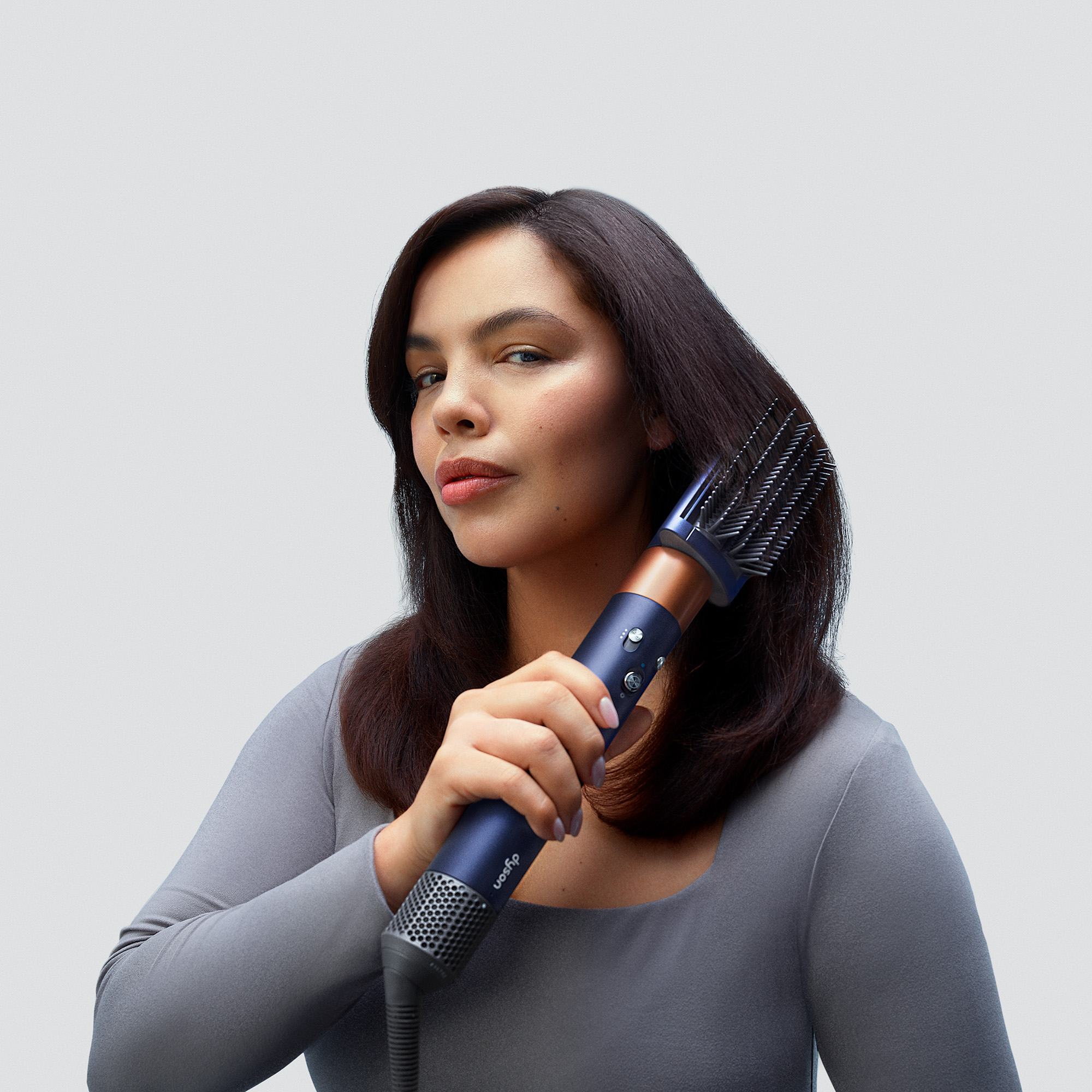 Eine Frau bürstet ihr Haar mit einer Stylingbürste. Die Bürste ist blau und kupferfarben. Der Hintergrund ist hellgrau.