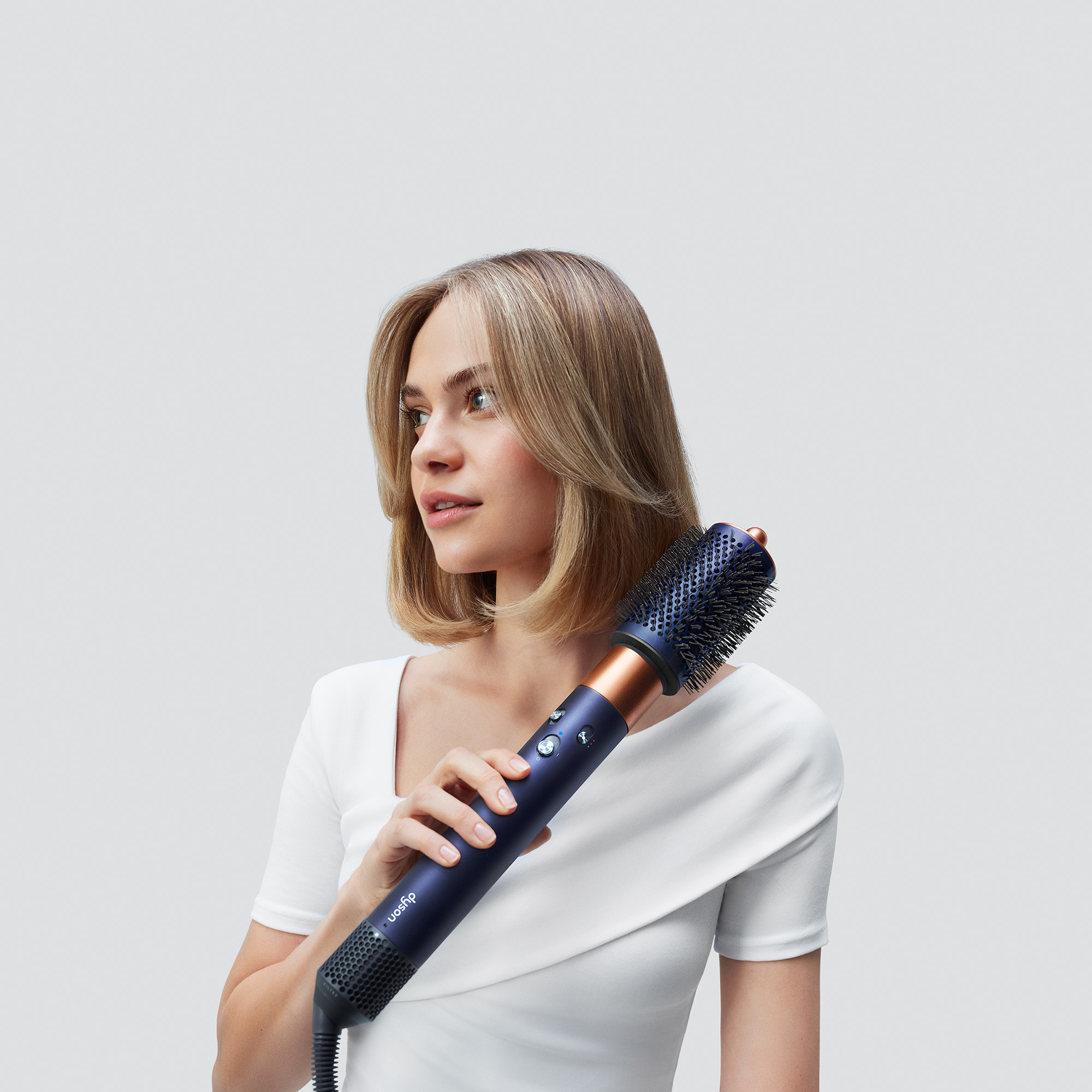Blonde Frau, die ihre Haare mit einem blauen und bronzefarbenen Dyson-Haarstyler stylt.