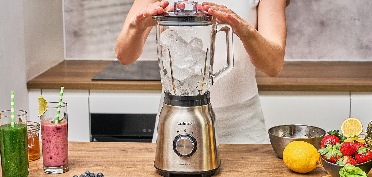 Osoba trzymająca blender z lodem, inne składniki na blacie.