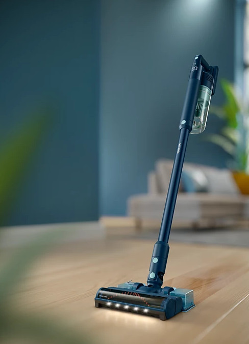 Odkurzacz bezprzewodowy w pokoju, oparty na drewnianej podłodze z niebieską ścianą w tle.