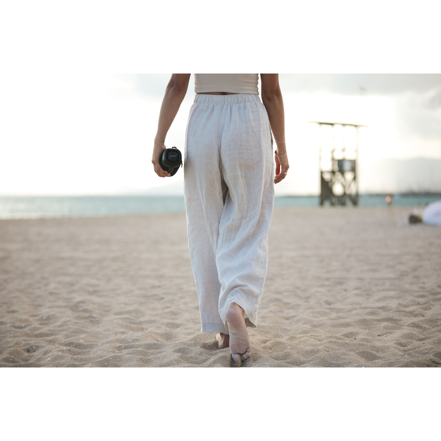 Persoon op strand, witte broek, houdt zwart object vast.