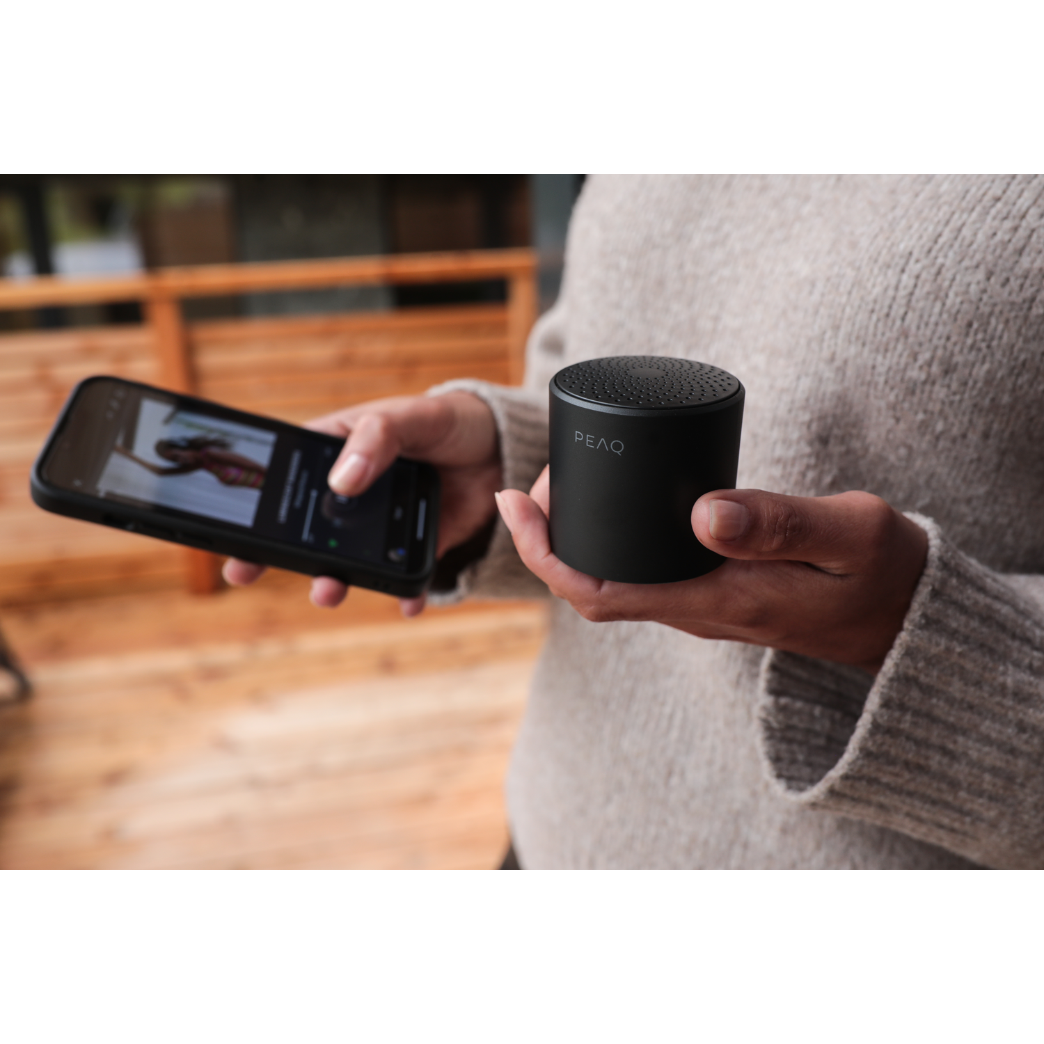 Persoon die een zwarte draagbare luidspreker en een smartphone vasthoudt met een muziek-app. Beige trui.
