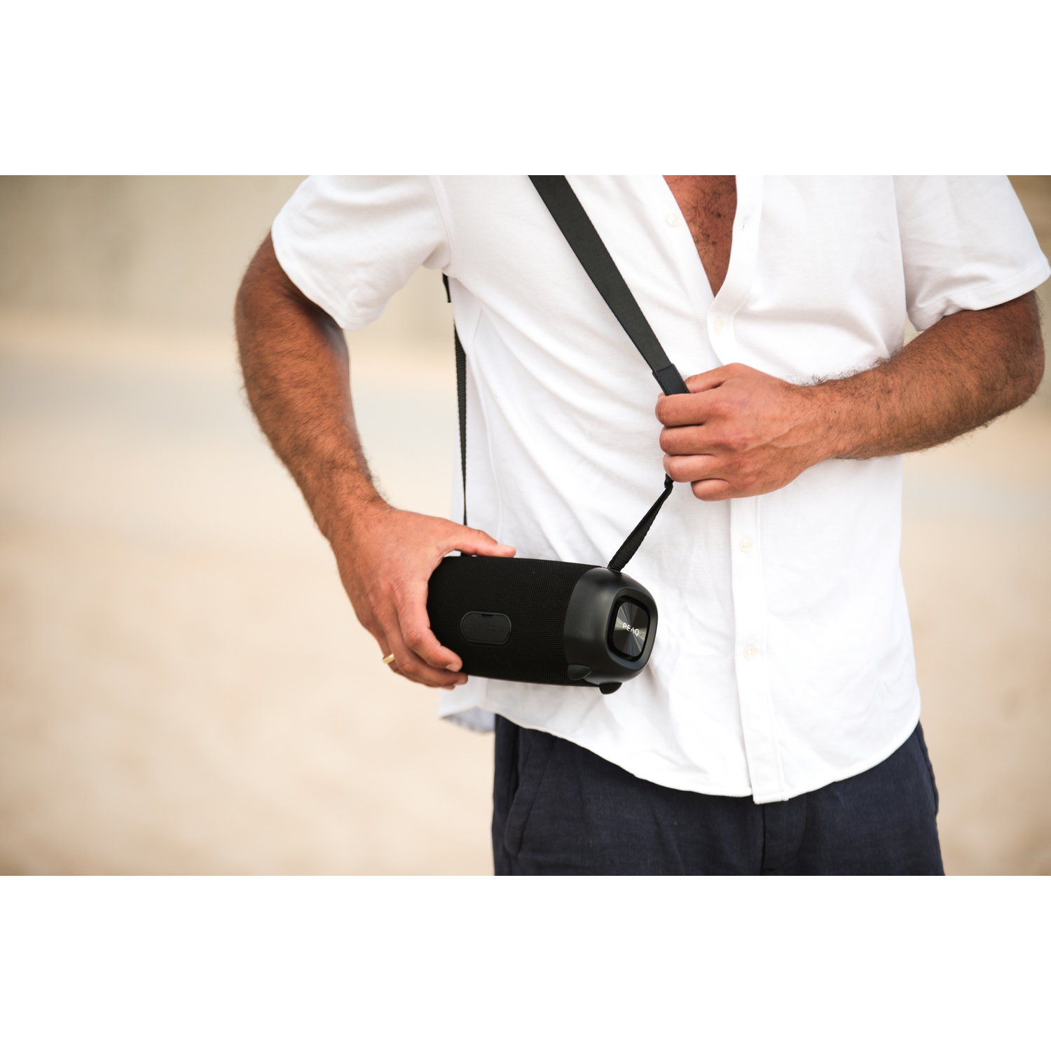 Man holding a black speaker with a black strap. He is in a white shirt, outdoors.
