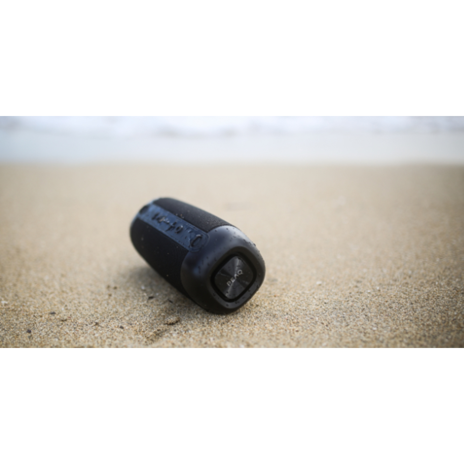 A black speaker on a sandy beach with the ocean in the blurred background.
