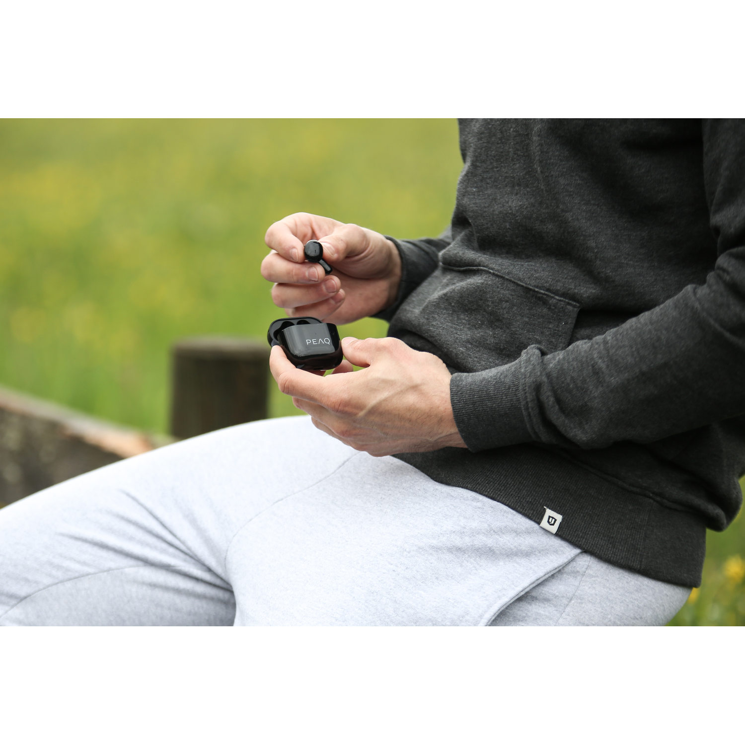 A person holding black earbuds in their hand, with the charging case. They are in a park.
