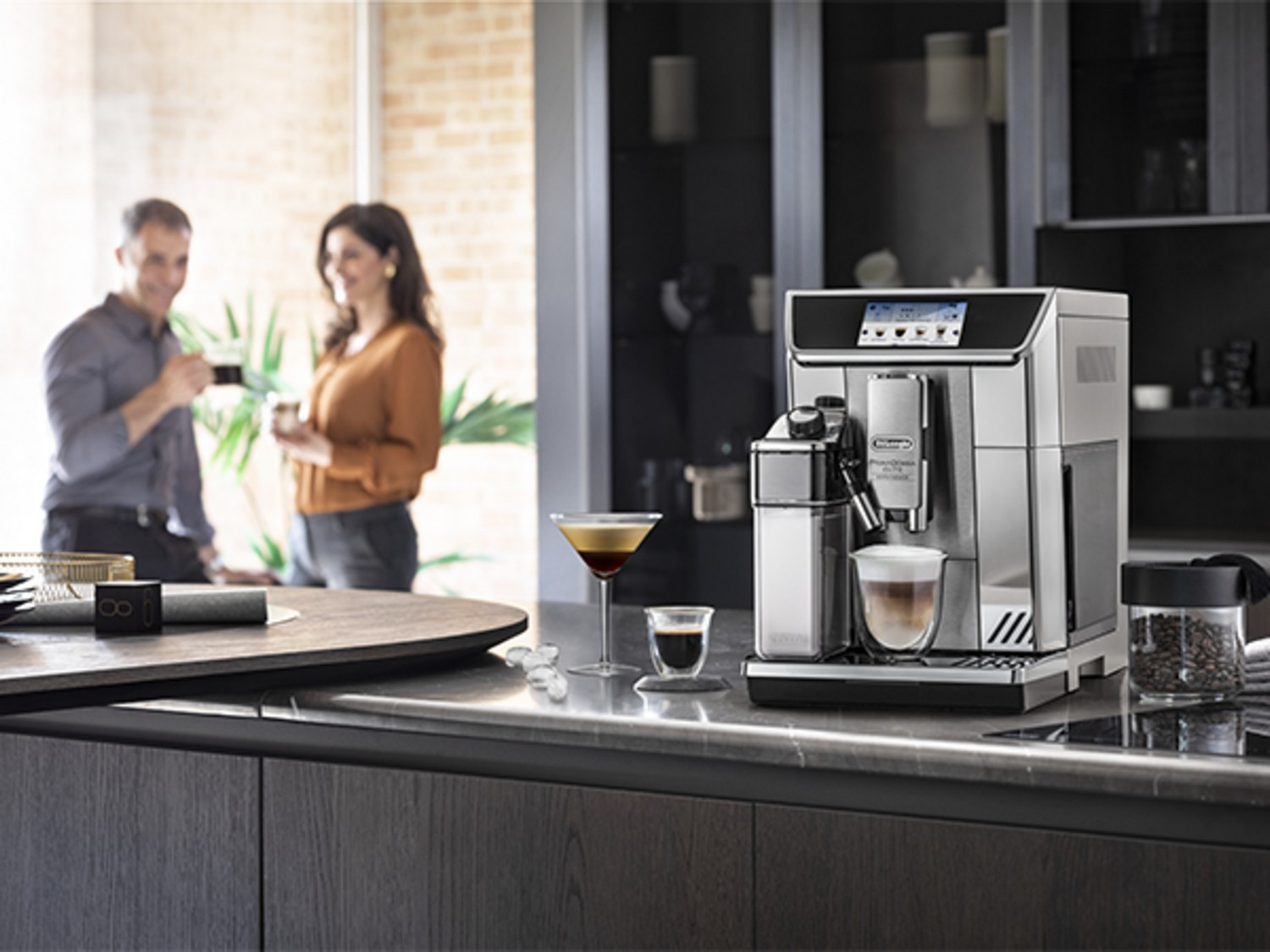 Una cocina moderna con cafetera, personas al fondo y copas en la encimera.
