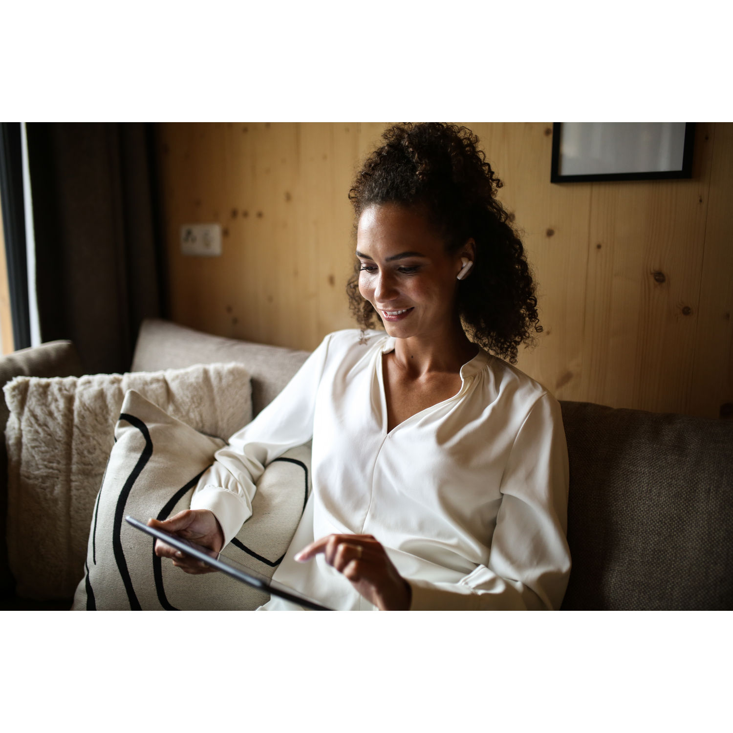 Woman on a sofa using a tablet. She is smiling, wearing a white shirt, and has earbuds in.