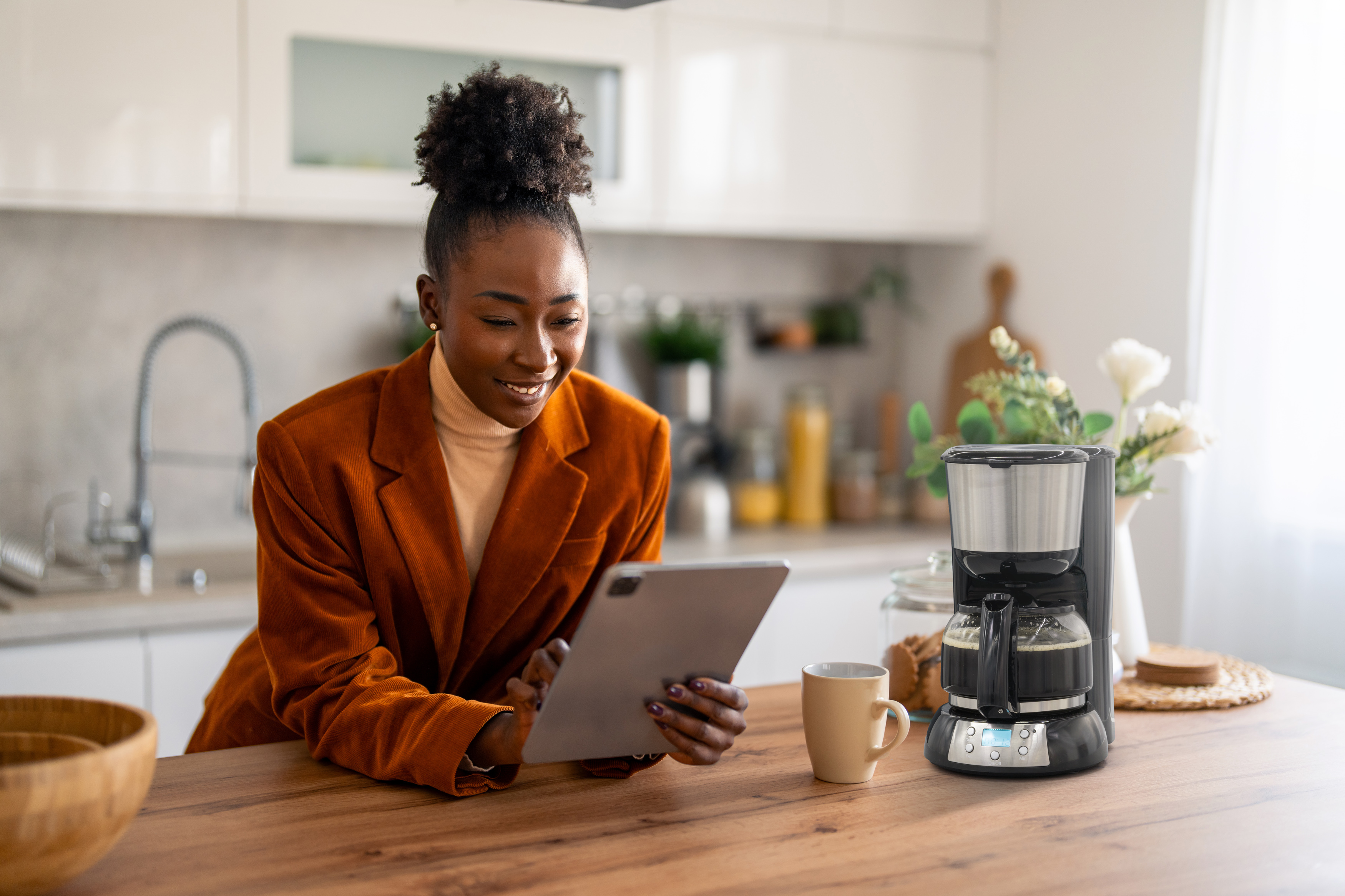 Frau in orangefarbenem Blazer benutzt Tablet in der Küche. Kaffeemaschine und Tasse auf dem Tisch.