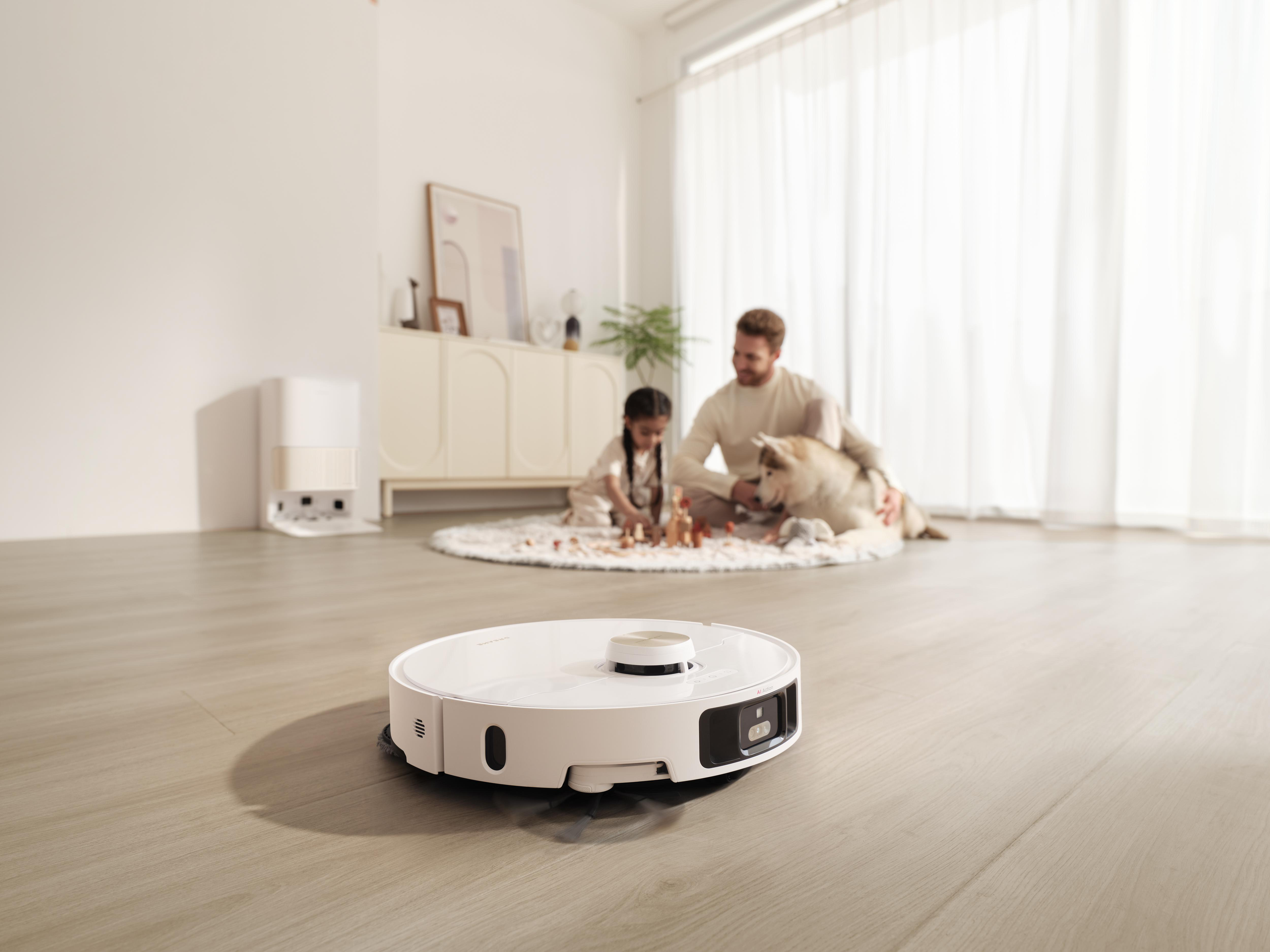 Ein weißer Roboter-Staubsauger auf Holzboden. Im Hintergrund eine Familie, mit Hund und Spiel.