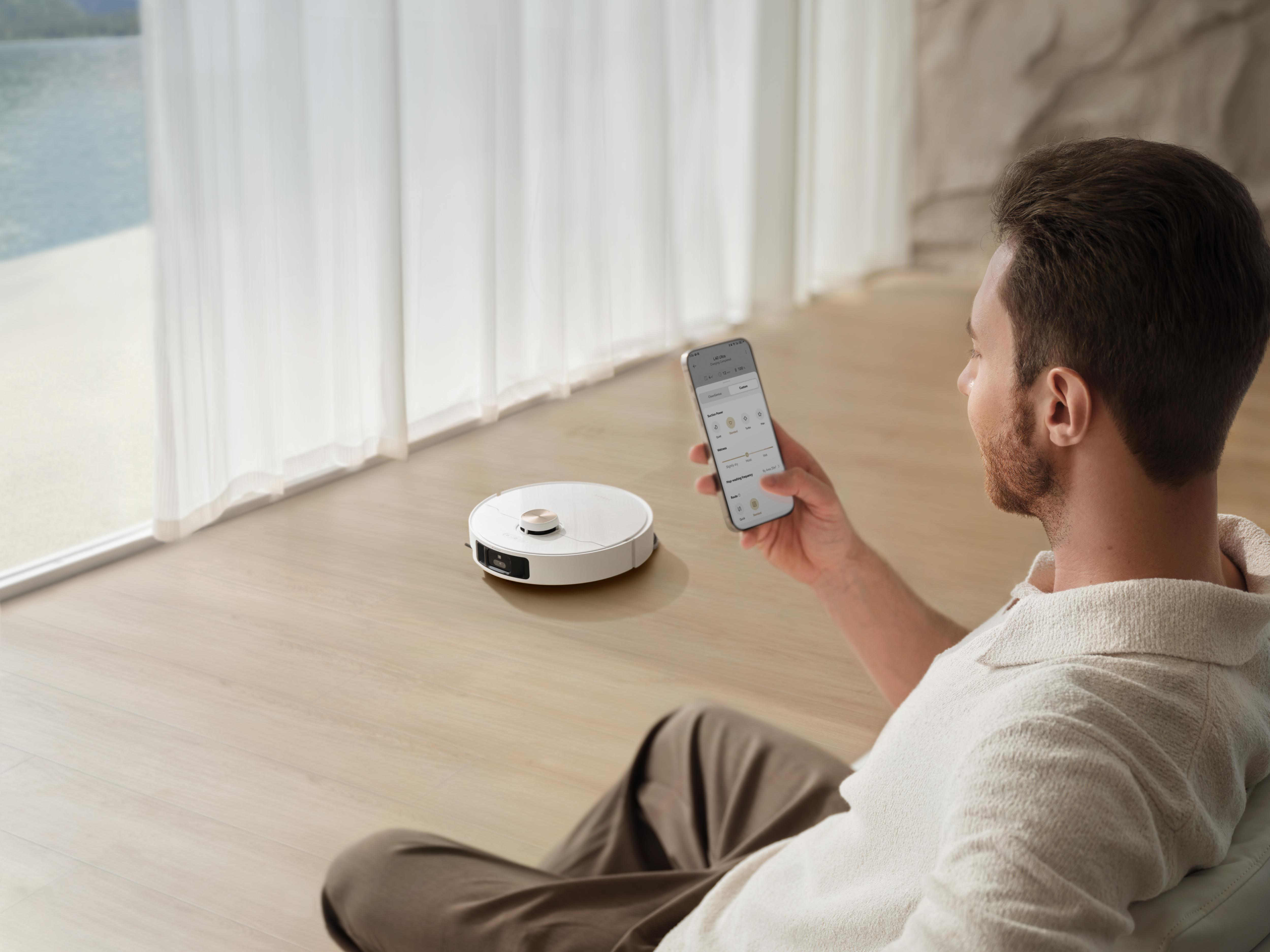Ein Mann hält ein Telefon in der Nähe eines Roboterstaubsaugers. Ein Fenster und ein Gewässer sind im Hintergrund.